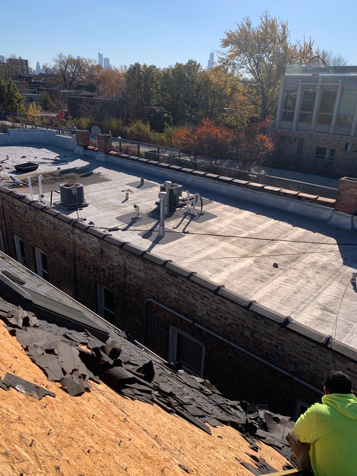 Rooftop with torn black membrane and orange debris, viewed from a sloped hillside beside a building.