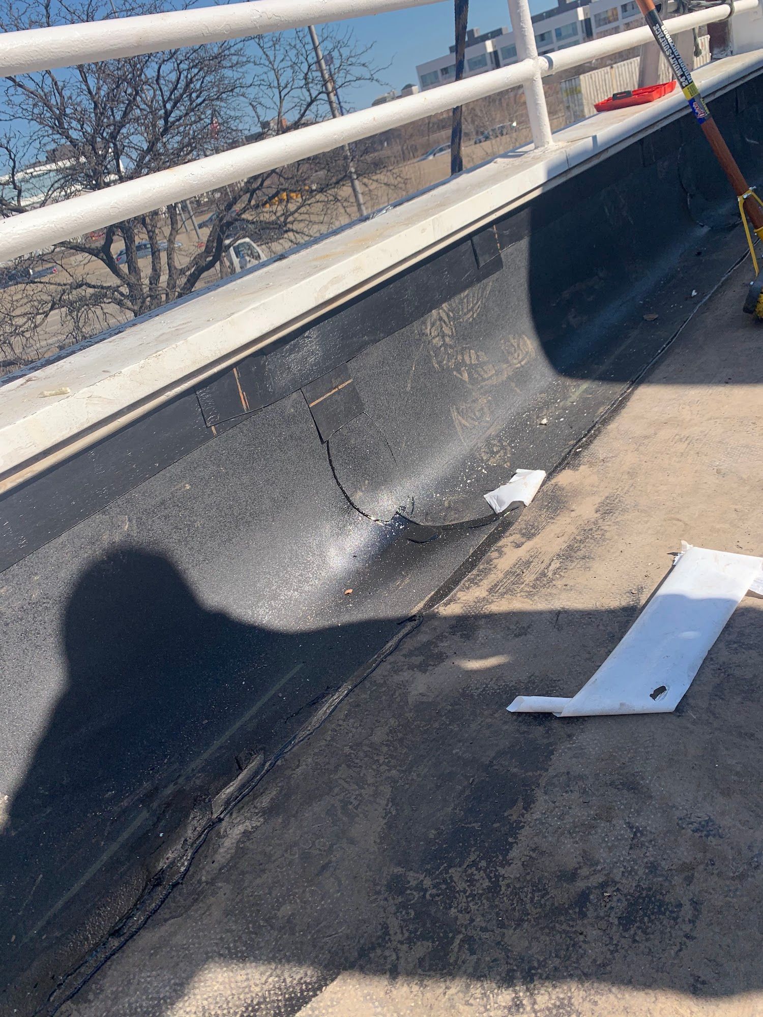 Damaged black roofing with a white metal piece and debris near a railing on a rooftop