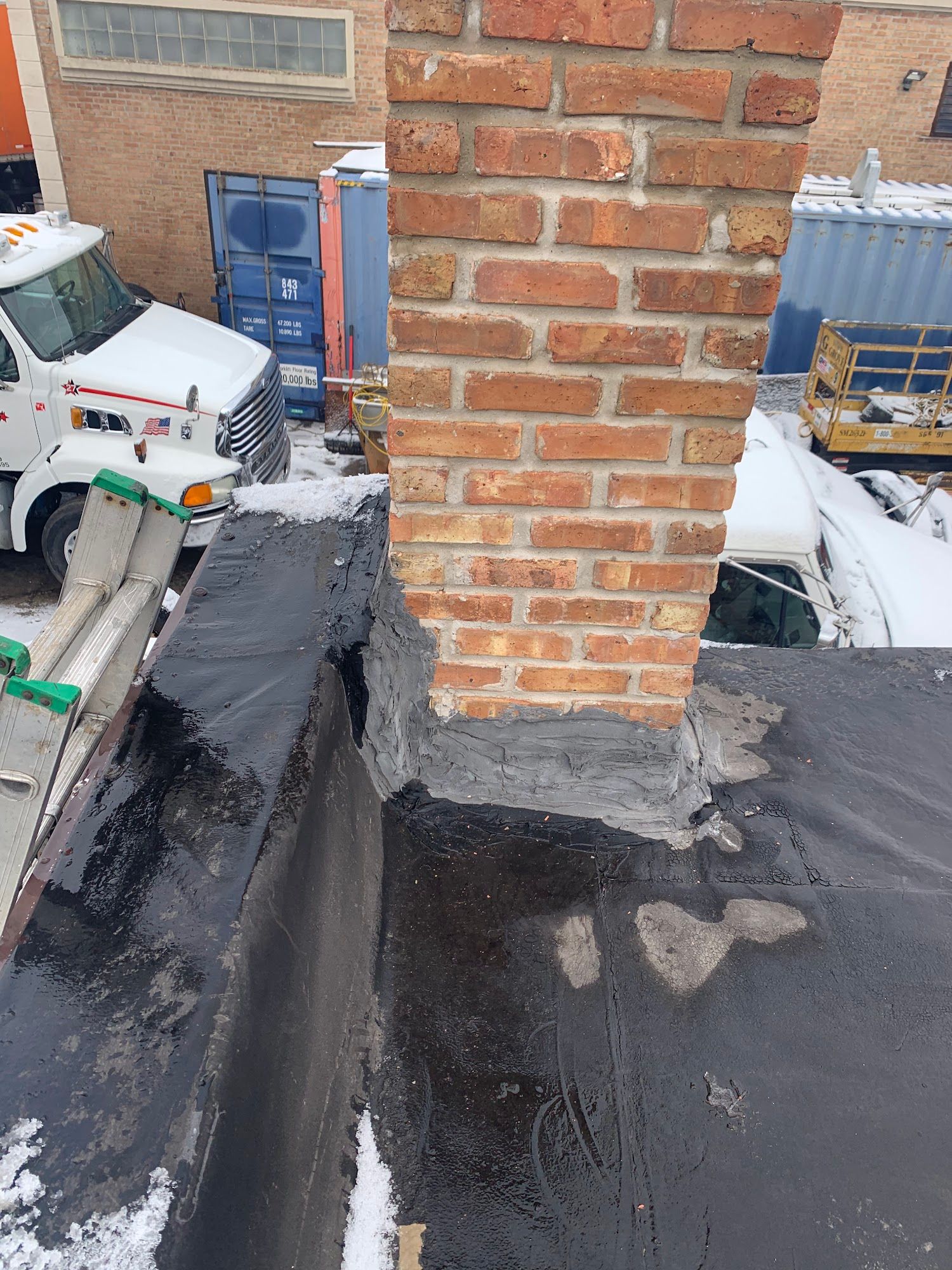 Snow-covered rooftop with a brick chimney and black waterproofing around the base.