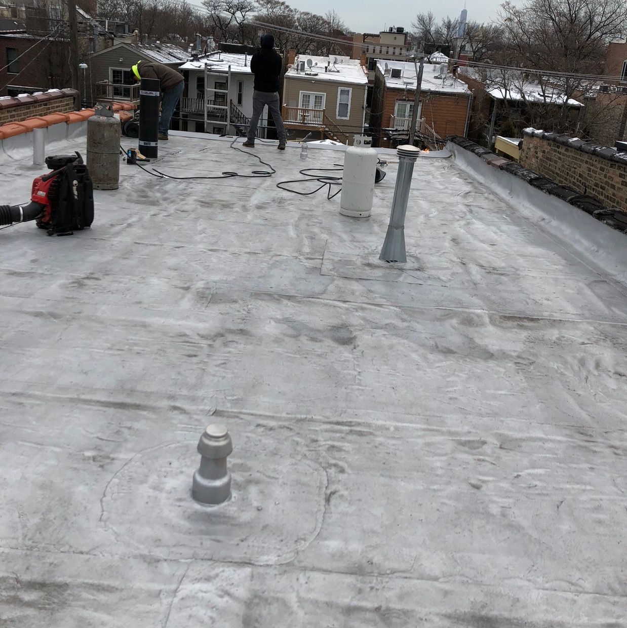 Snow-covered rooftop with two workers and roofing equipment near a city skyline