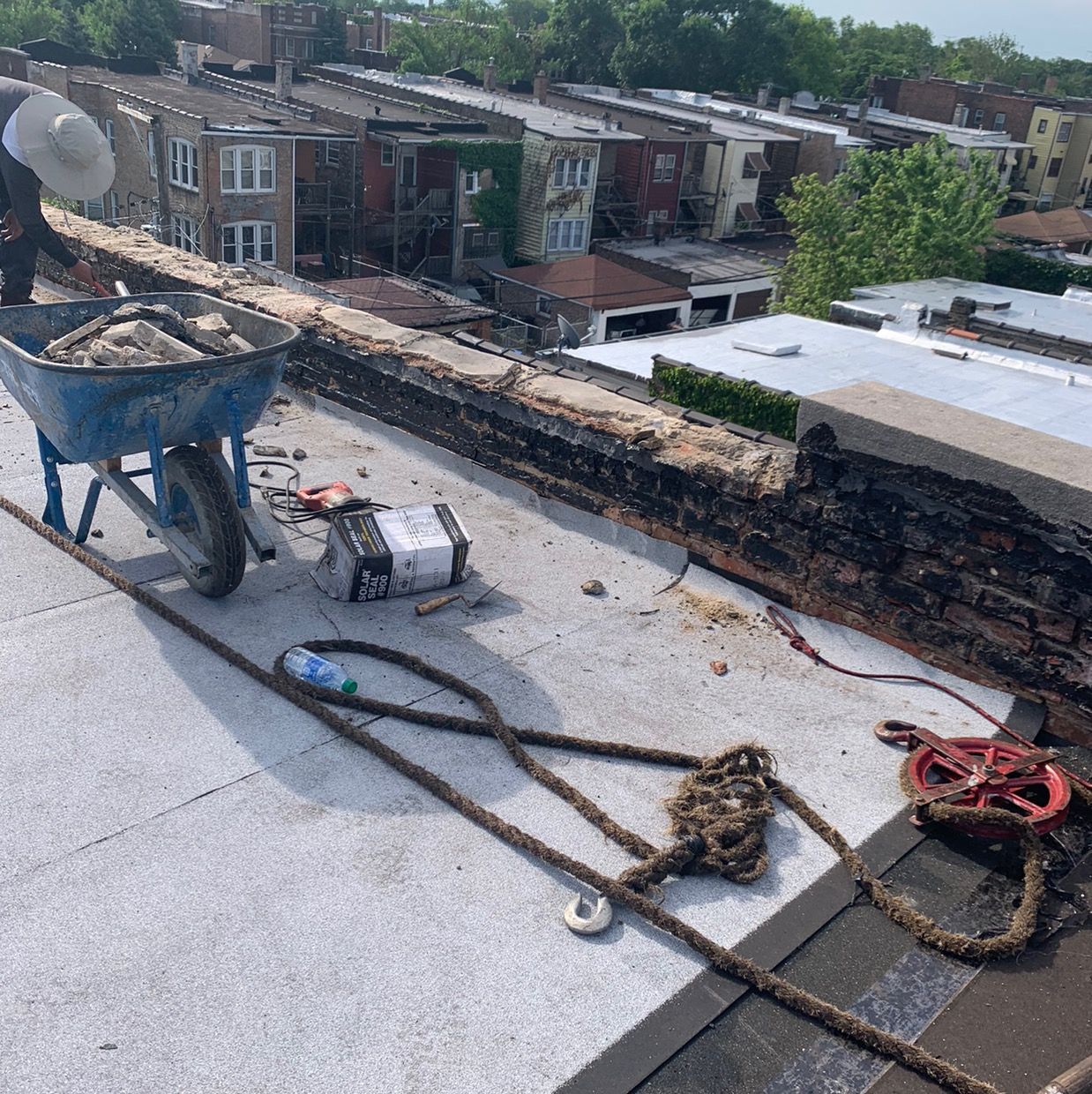 Rooftop with wheelbarrow, ropes, and tools beside a damaged brick row of buildings in the background