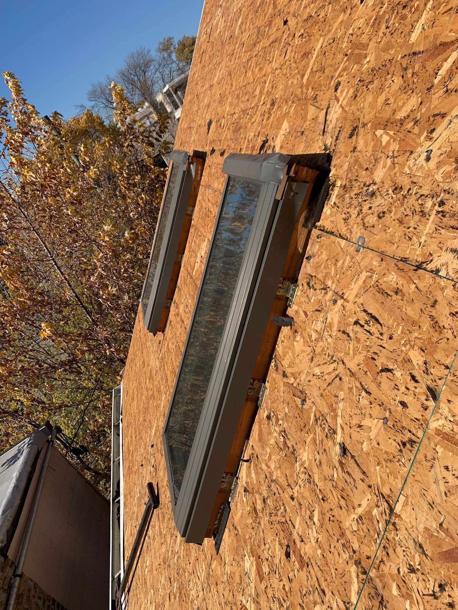 Sideways view of a house roof with a skylight and trees against a blue sky.