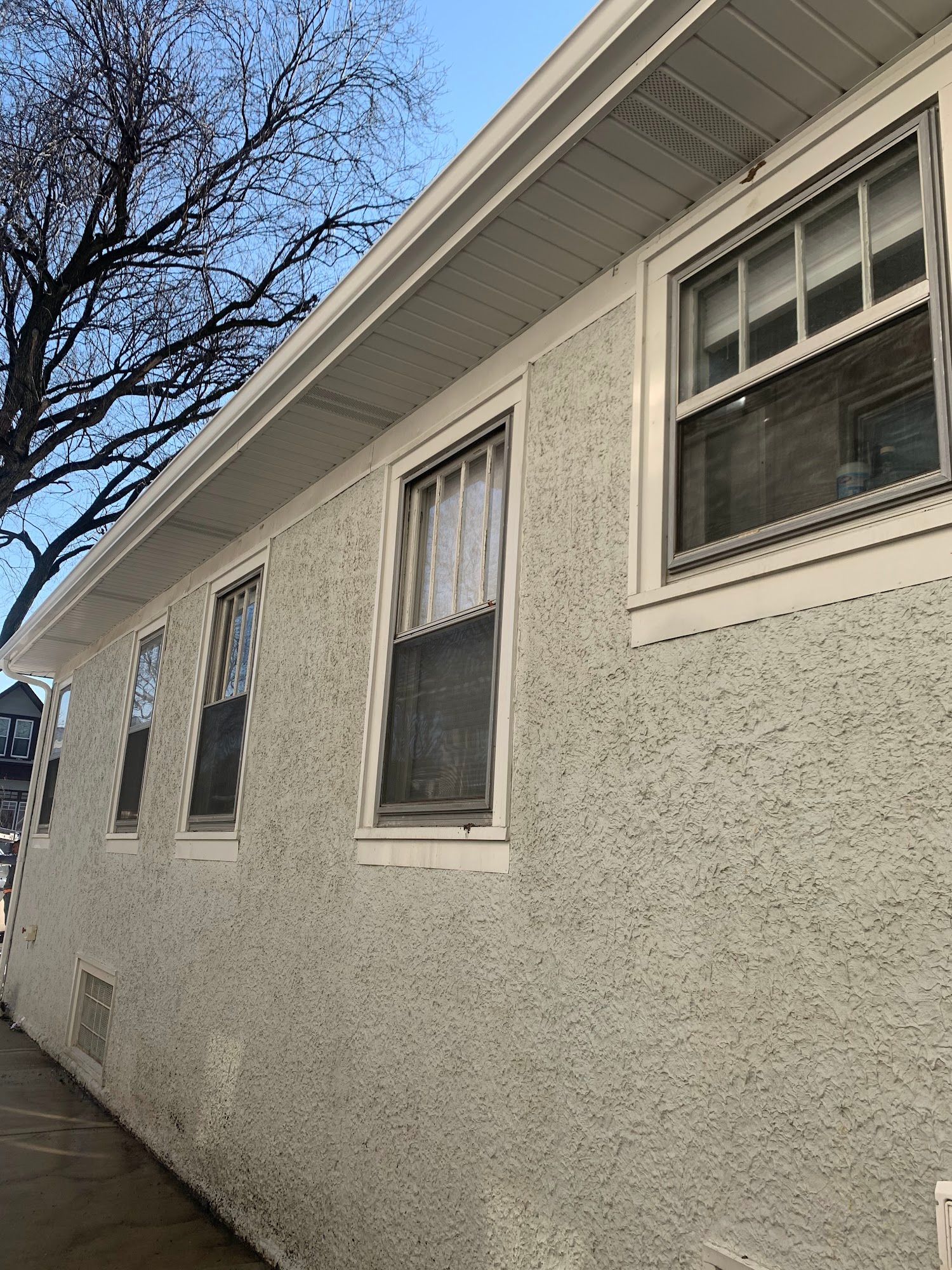 White stucco building exterior with several windows along a sidewalk, under a bare tree.