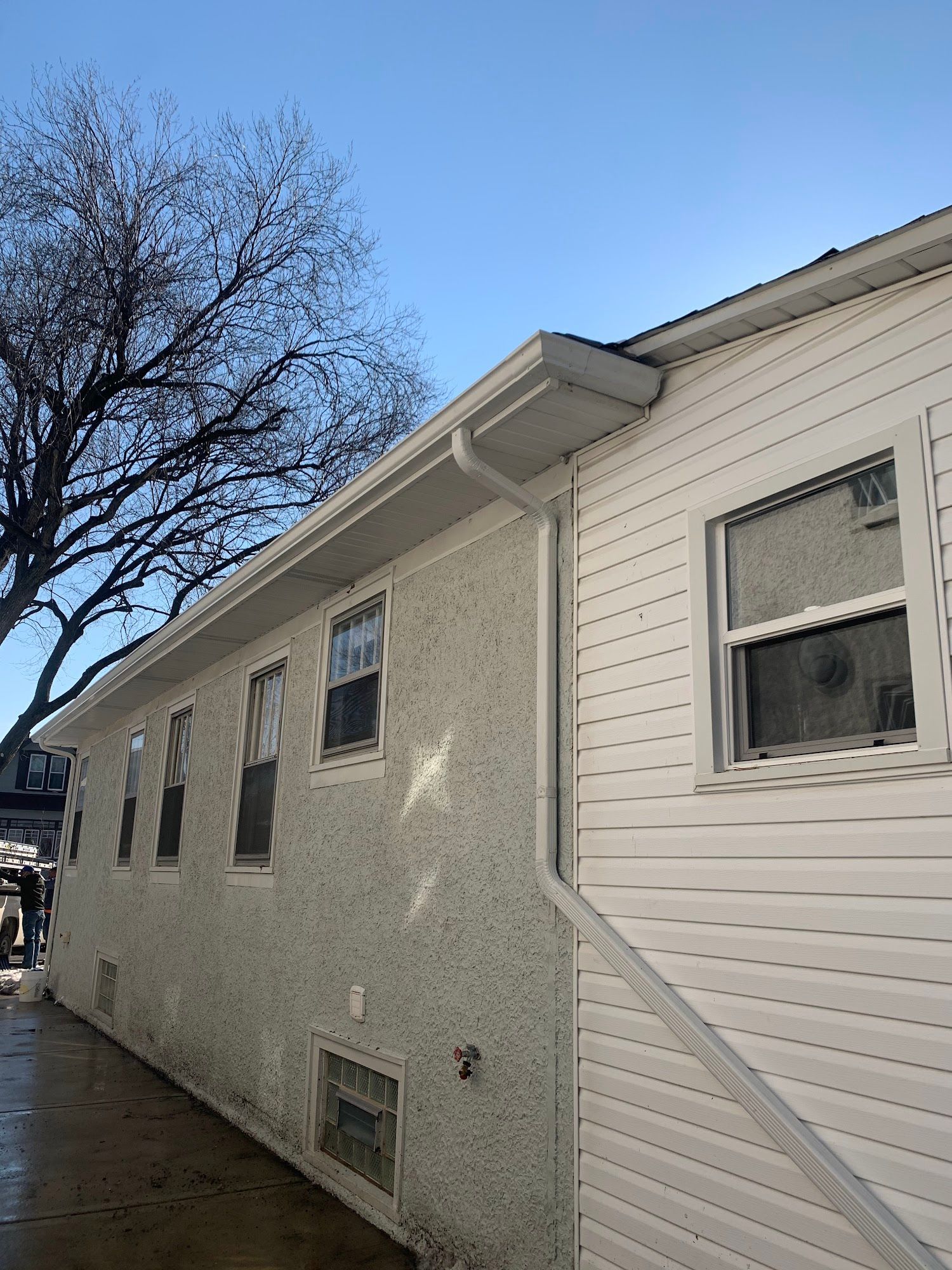 Side view of a white house with vinyl siding beside a wet driveway and leafless tree under a blue sky
