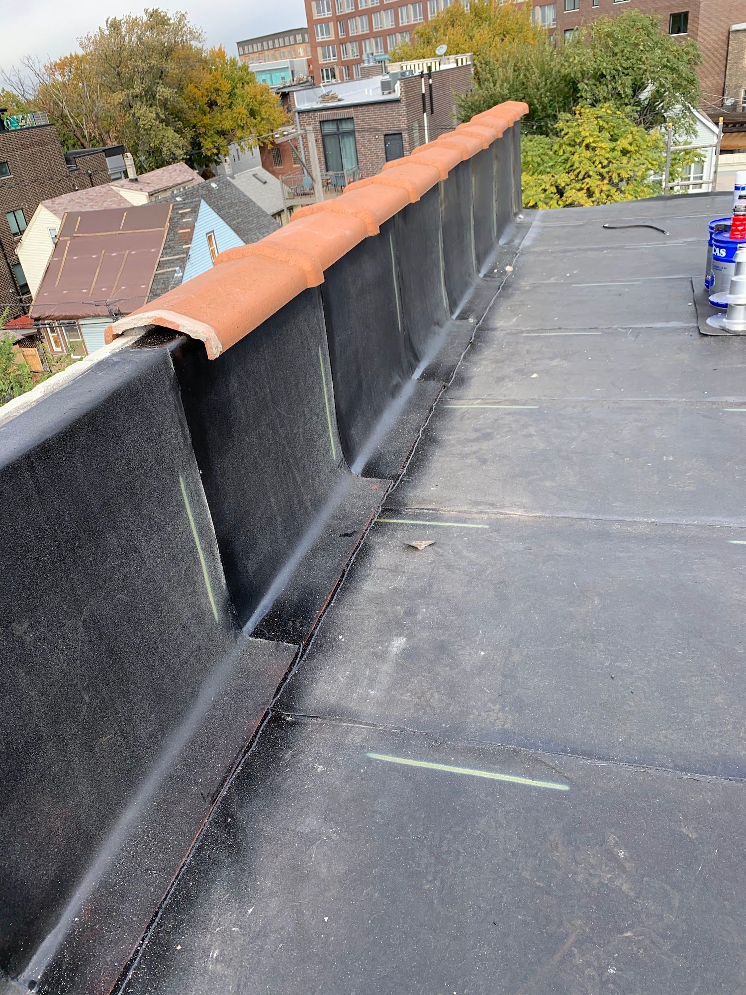Rooftop with black surface and low parapet wall capped in orange, overlooking trees and buildings.
