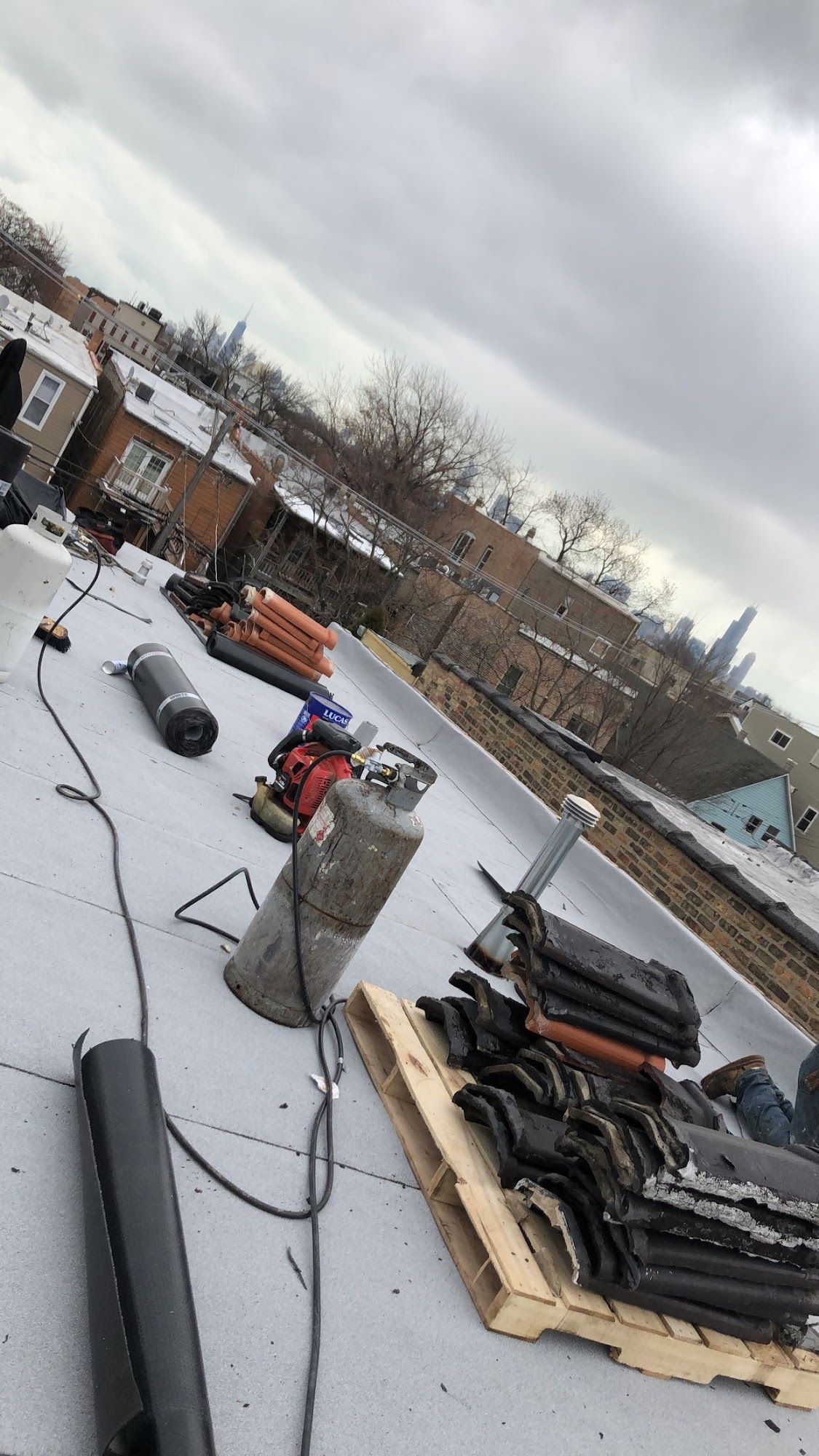 Snow-covered rooftop with stacked black materials, cables, and a worker in red near a brick building.