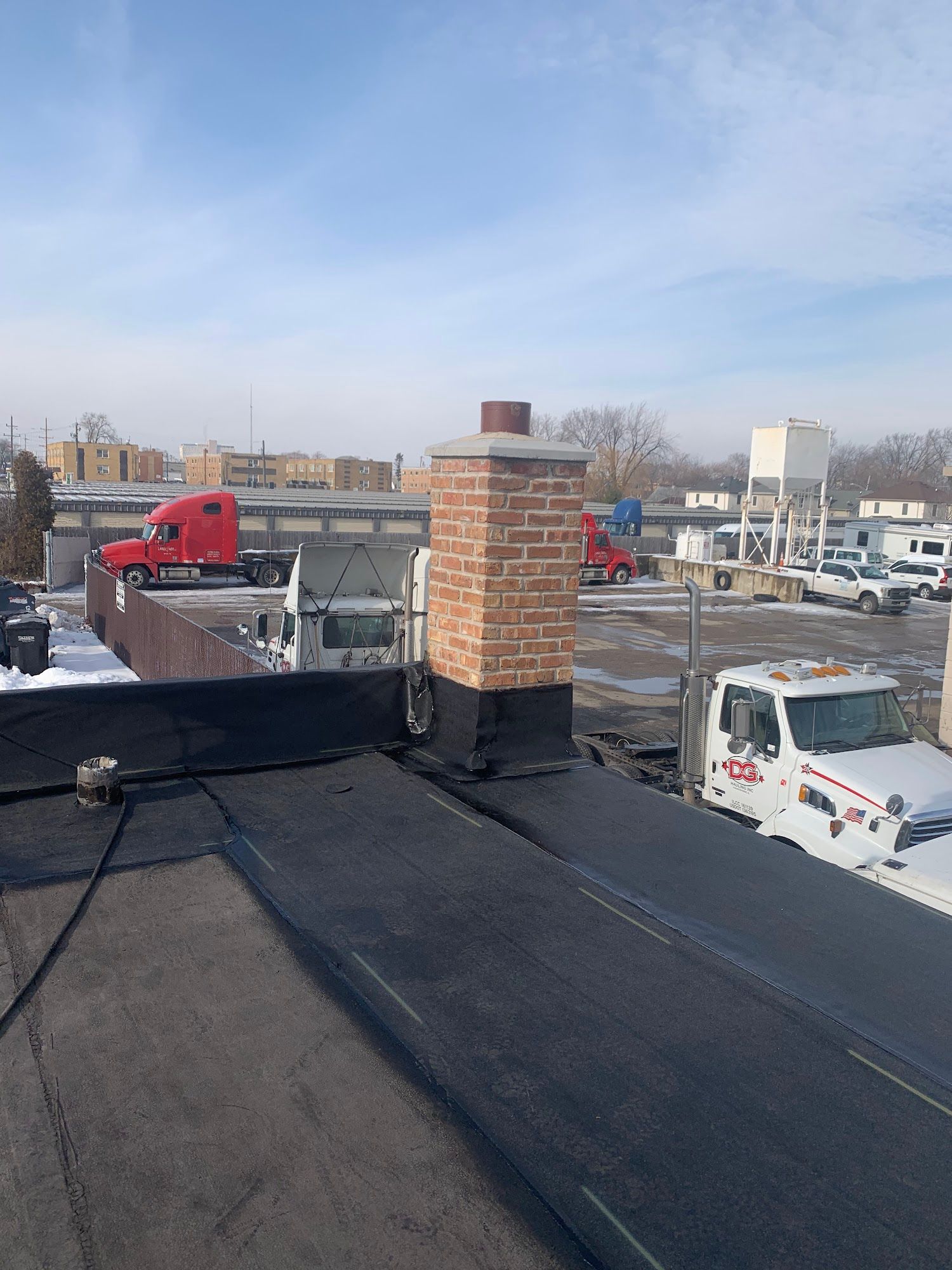Snowy rooftop with a brick chimney, parked trucks, and city buildings under a clear sky