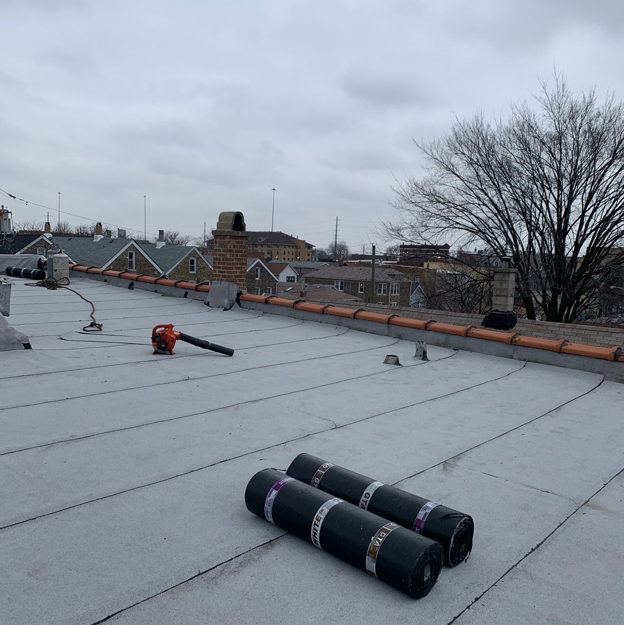 Flat rooftop with rolled roofing materials, an orange tool, and cloudy sky over nearby houses.