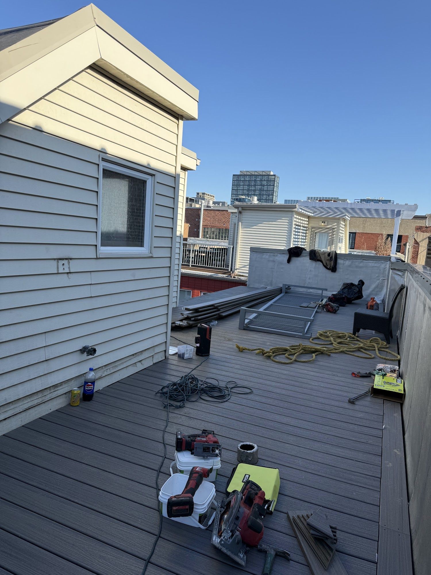 Rooftop construction site with tools, tarps, and materials beside a white building under a clear sky