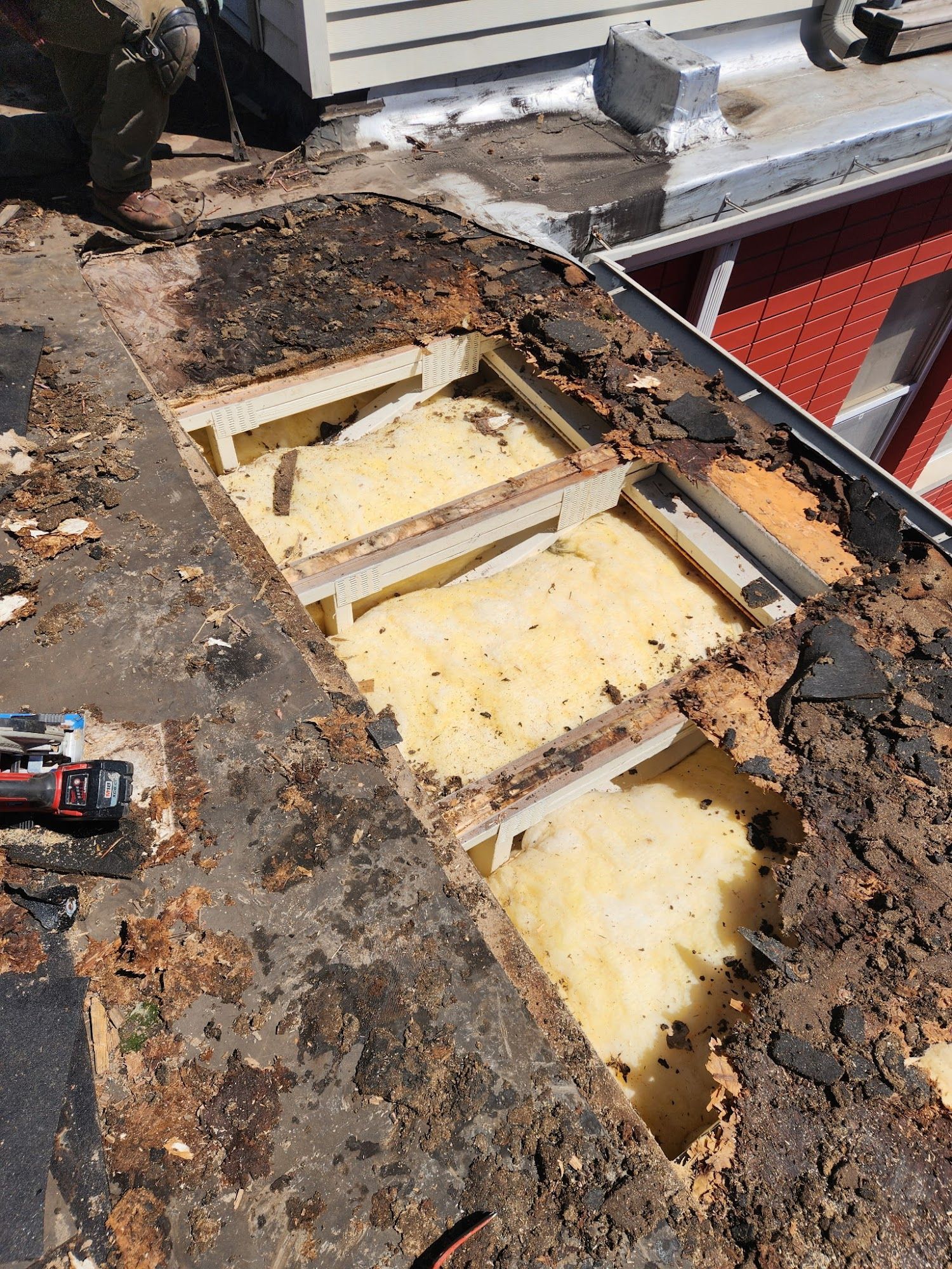 Rooftop under repair with exposed insulation, wooden framing, and scattered debris near a red building.