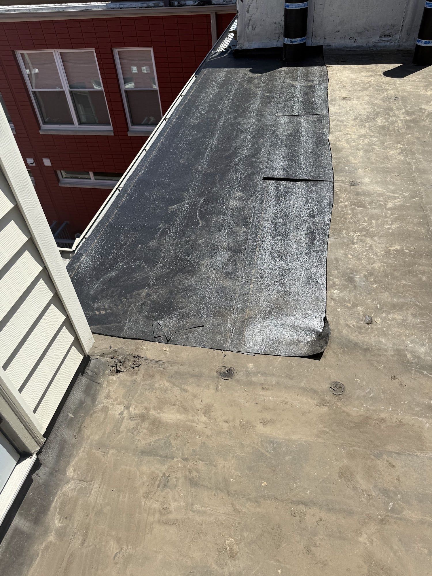 Rooftop with a black sloped roof section beside a tan flat roof and a red brick building in the background