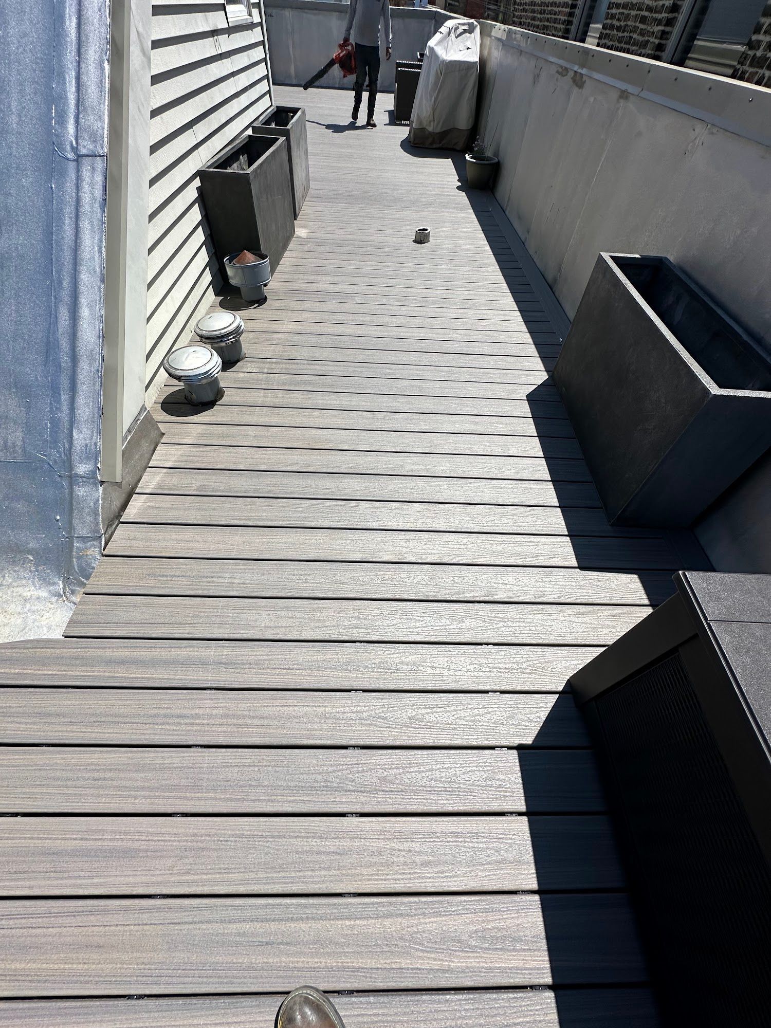 Narrow wooden boardwalk between a building and a concrete wall, with planters and a person in the distance.