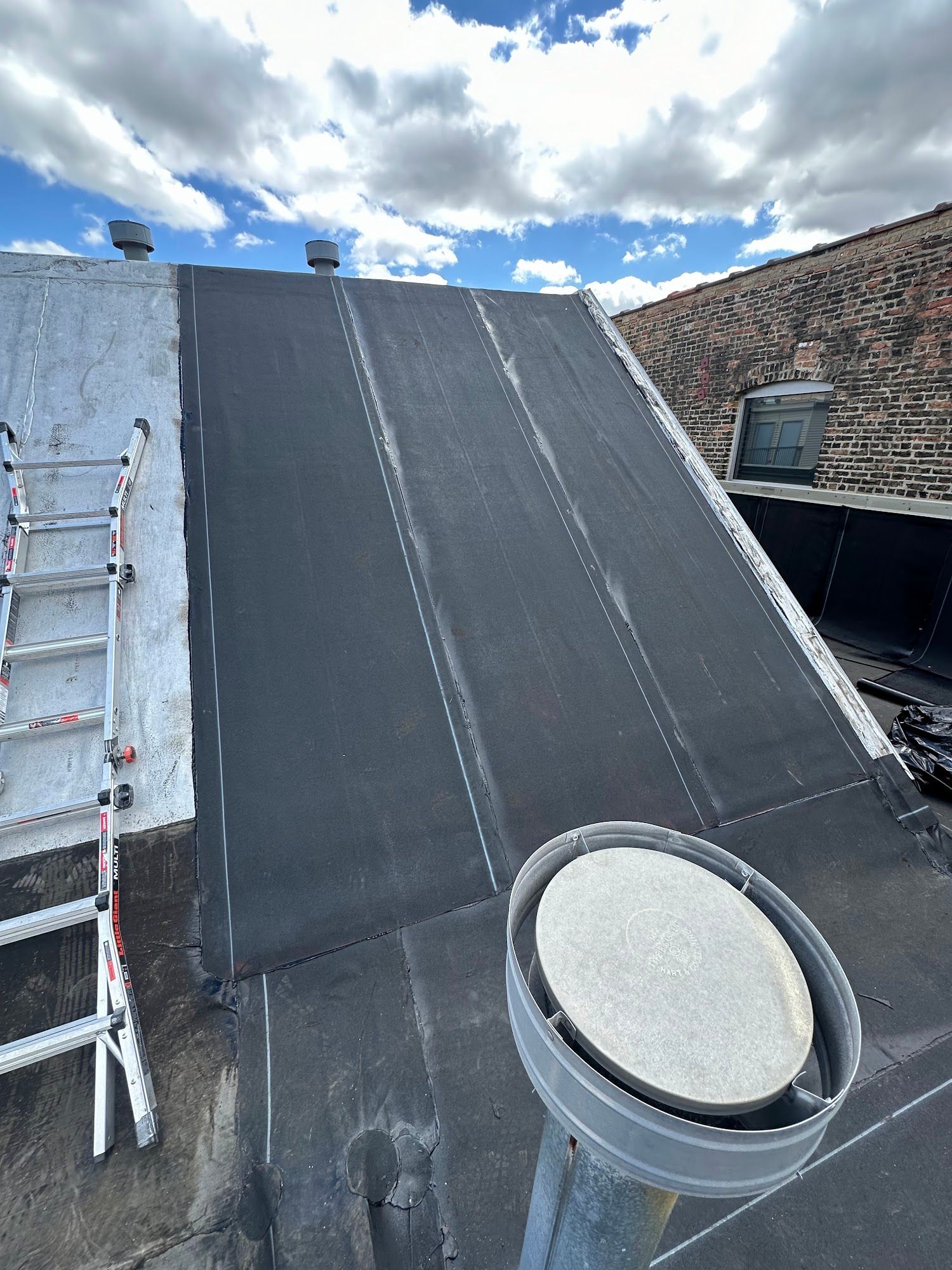 Rooftop with a black sloped roof membrane, ladder on the left, and a round vent in the foreground under a cloudy sky