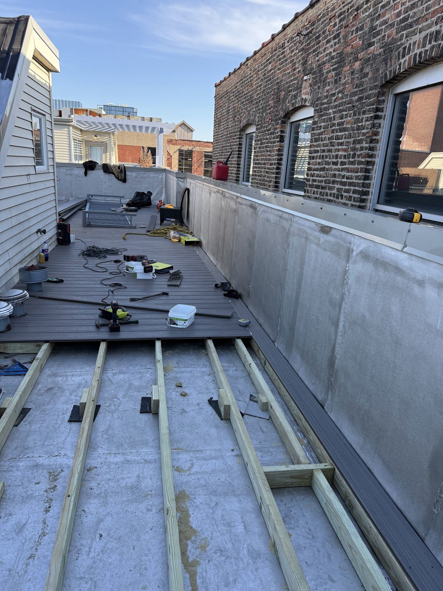 Rooftop walkway under construction with boards, metal rails, tools, and brick buildings on both sides