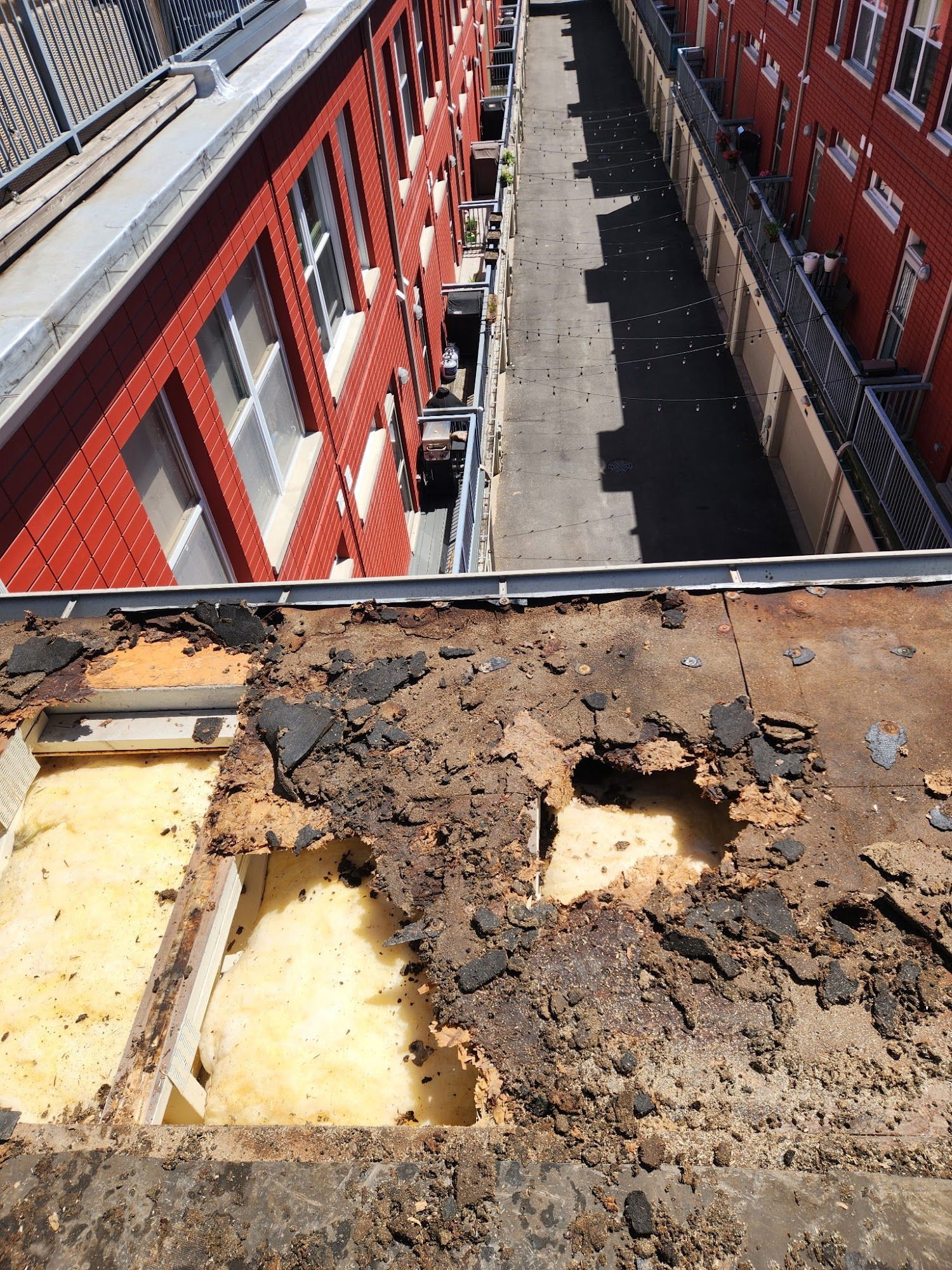 Rooftop view over a narrow alley between red brick buildings; foreground shows damaged, crumbling roof material.