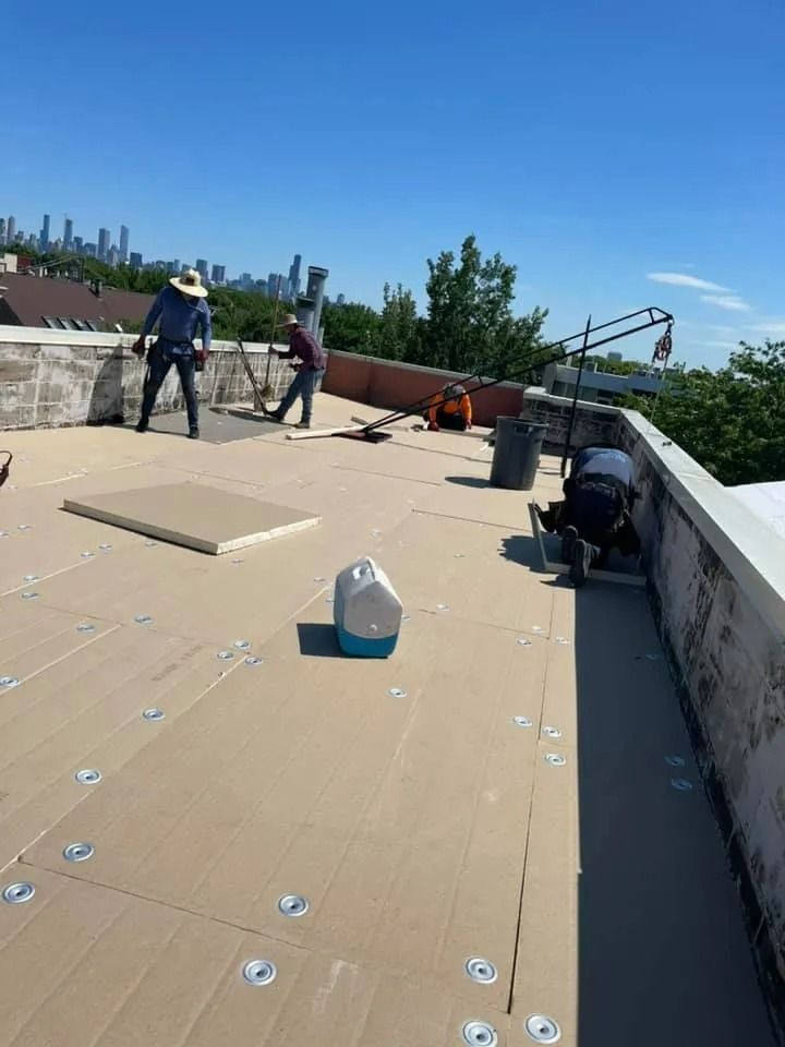 Rooftop worksite with two workers, tools, and a white jug on a tan covered roof under a clear blue sky