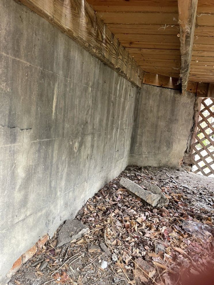 Concrete wall under a deck with leaf-covered ground and a small broken concrete block.
