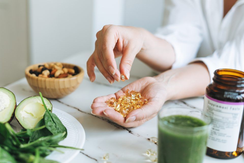 Hands holding supplements beside a green smoothie, capsules, and a jar on a bright kitchen counter