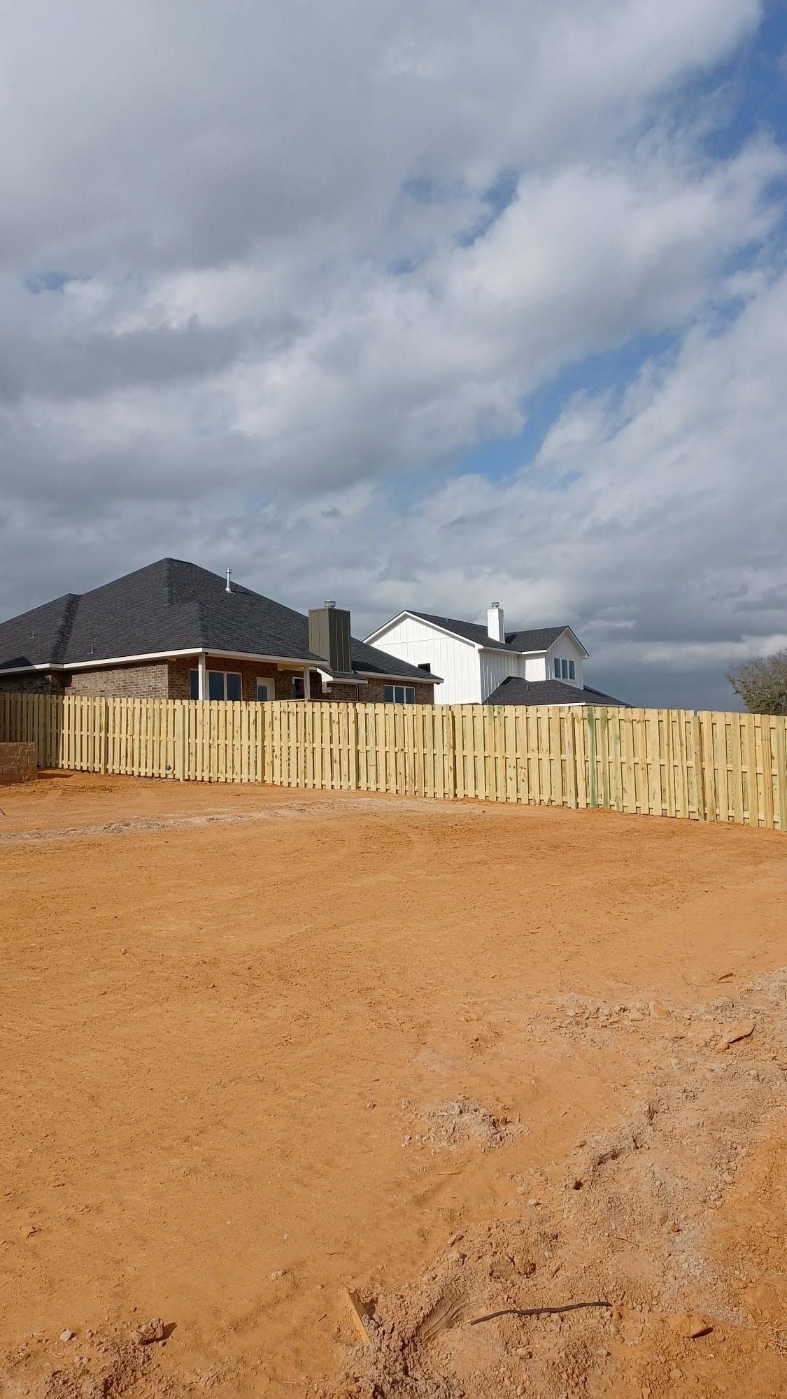 Dirt lot with a wooden fence and suburban houses under a cloudy sky