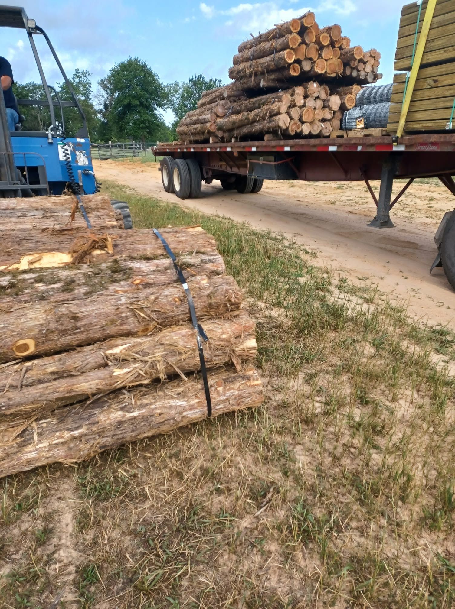 Logs stacked beside a flatbed trailer loaded with cut timber on a dirt road, with a blue machine nearby.