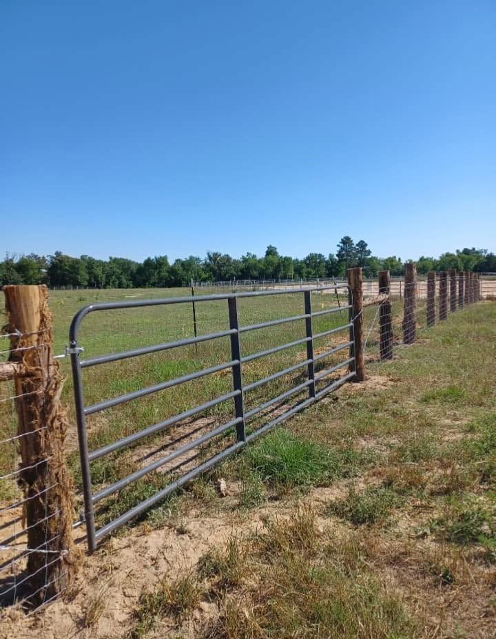 Metal farm gate between wooden fence posts beside a dry grassy field under a clear blue sky