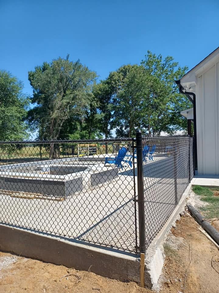 Chain-link fence enclosing raised concrete pads beside a white building, with blue playground equipment and trees behind.