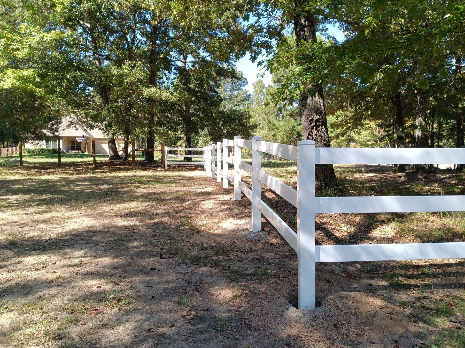 White fence along a shaded dirt path in a wooded park near a small building