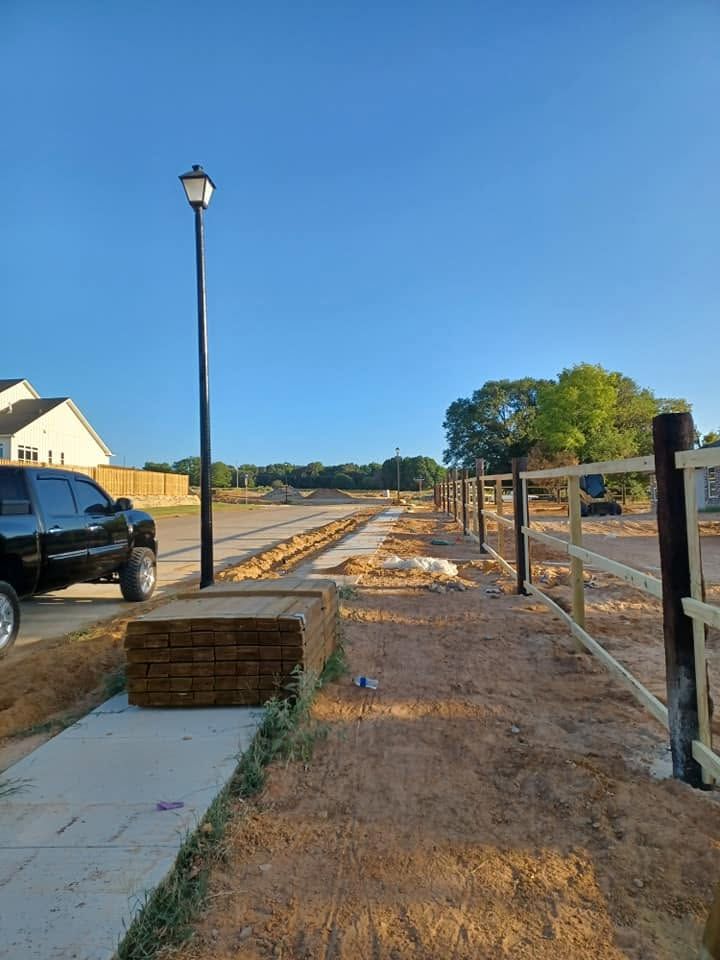 New suburban sidewalk under construction beside a street, with lamp posts, dirt, and parked vehicles