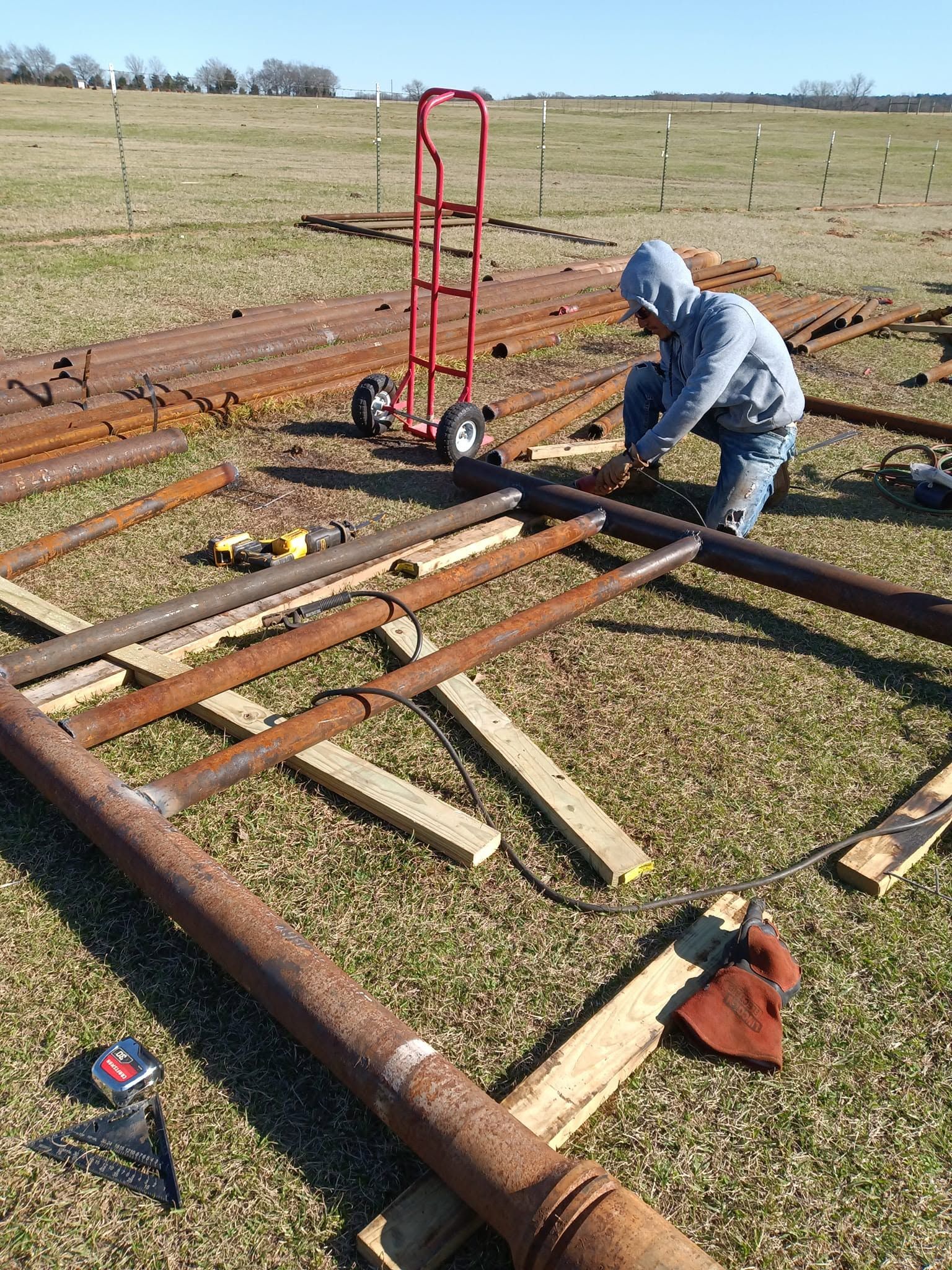 Person kneeling by metal gate frame in a field, using tools beside a red hand truck.