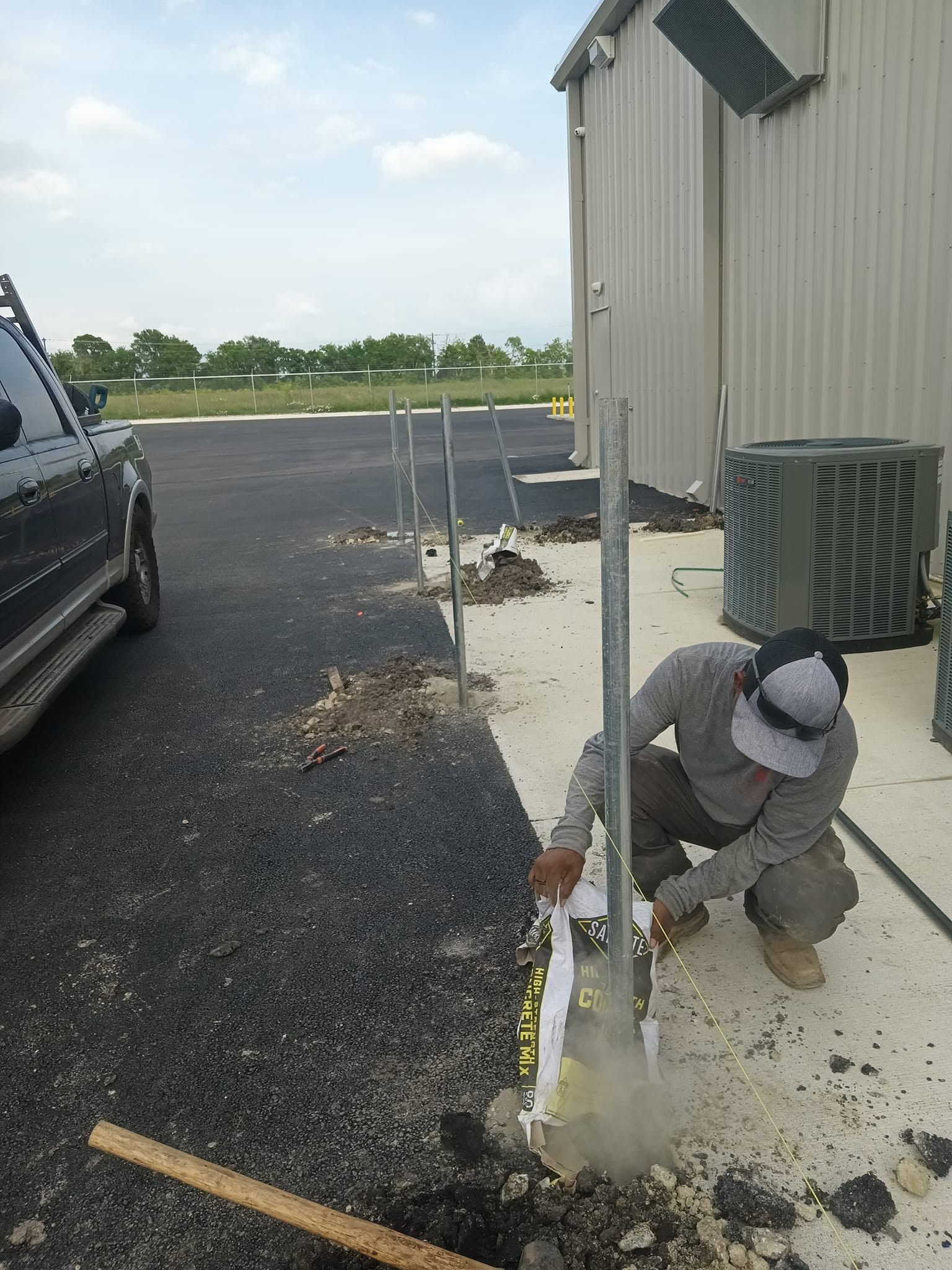 Worker installing metal post near building and HVAC unit on gravel beside parking lot.