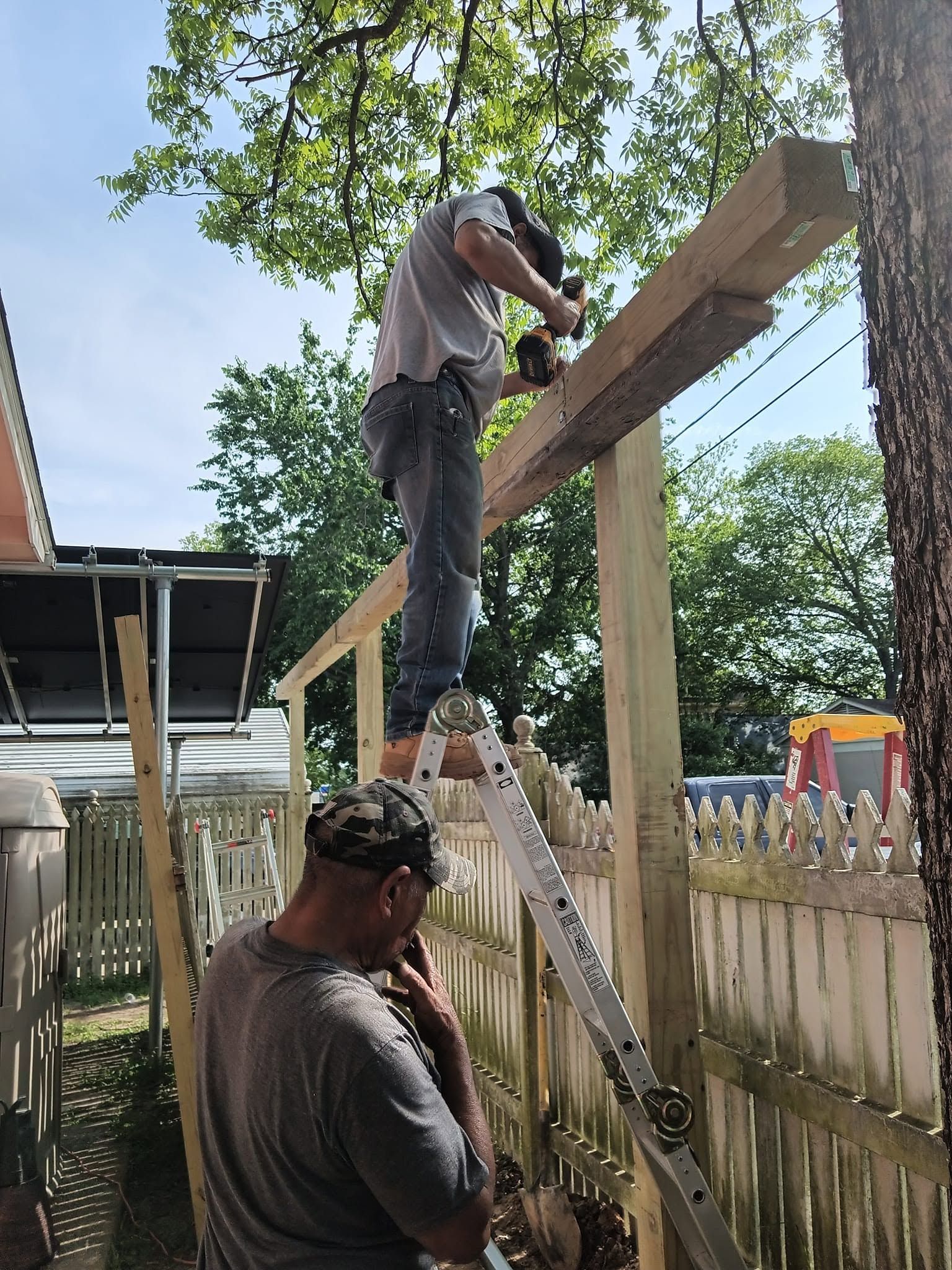 Two workers install a wooden railing on a raised deck beside a picket fence.