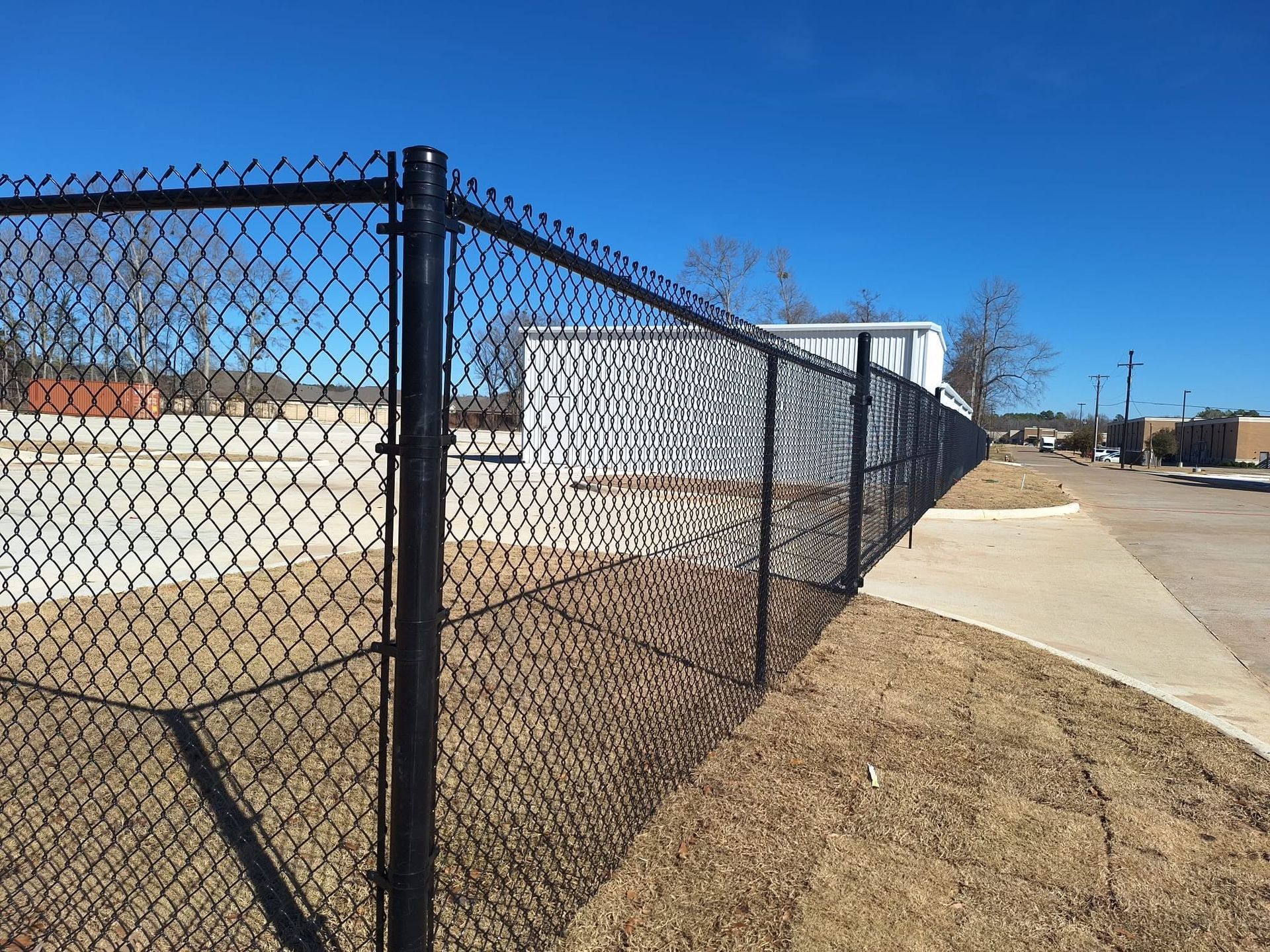 Chain-link fence beside a sidewalk, with a grassy slope and industrial buildings under a blue sky.