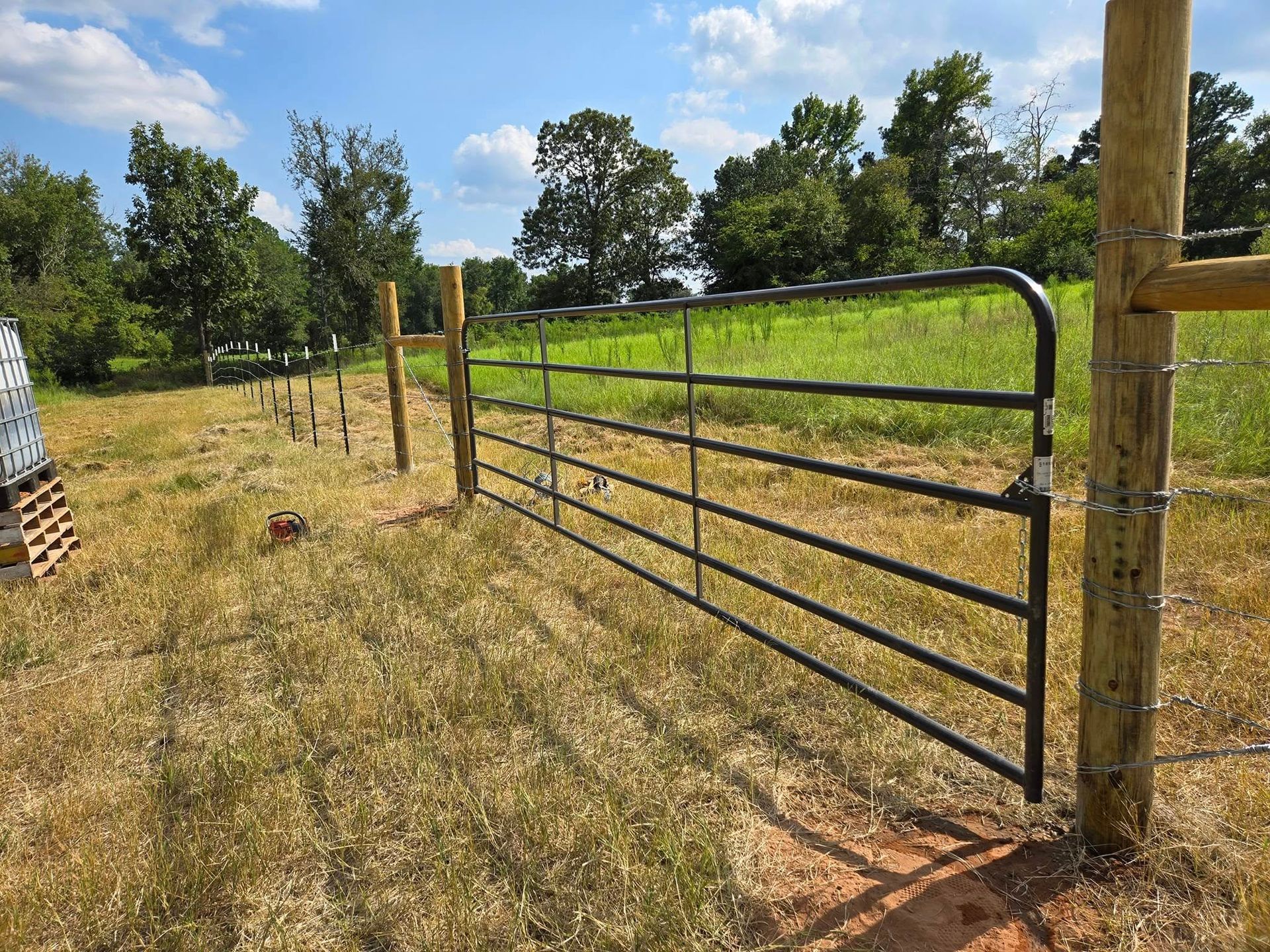 Black metal farm gate beside a wooden post along a grassy rural fence line under a blue sky.