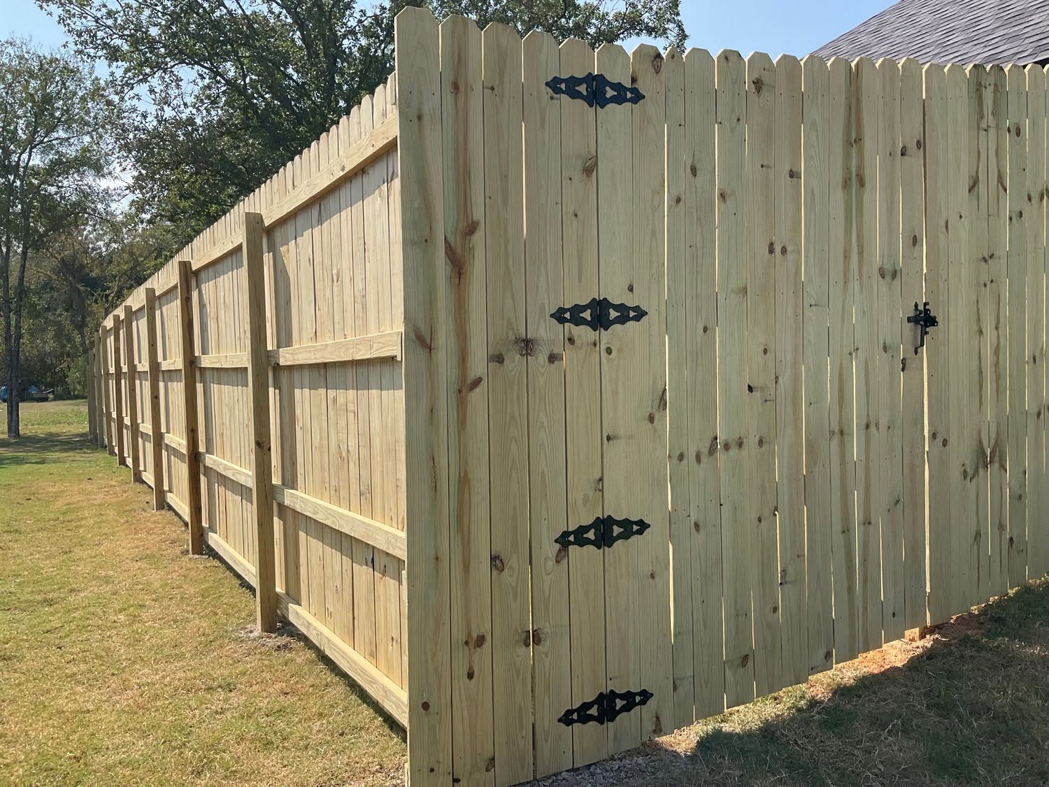 Wooden privacy fence with black metal hinges in a grassy yard under trees