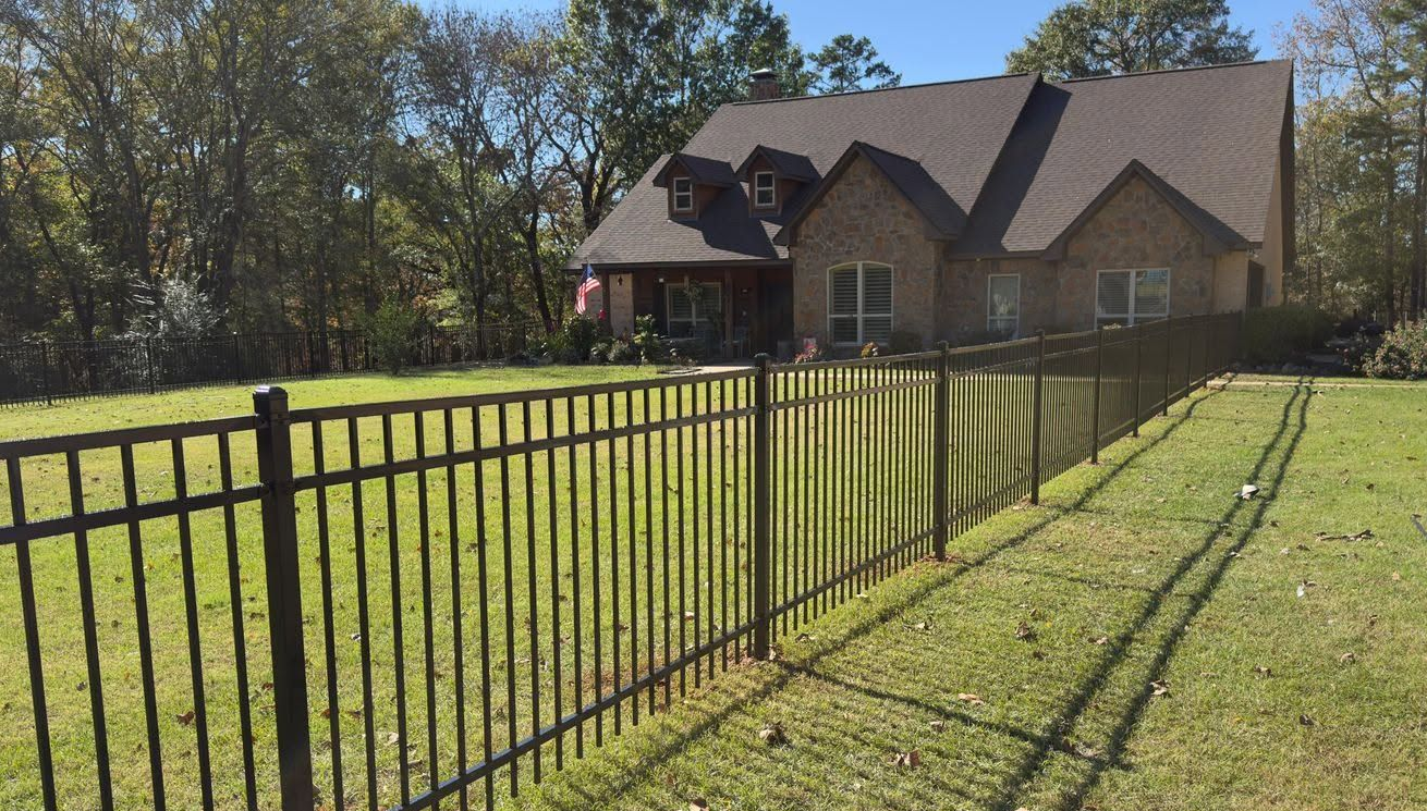 Brick house behind black metal fence on a sunny grassy lawn, with trees in the background