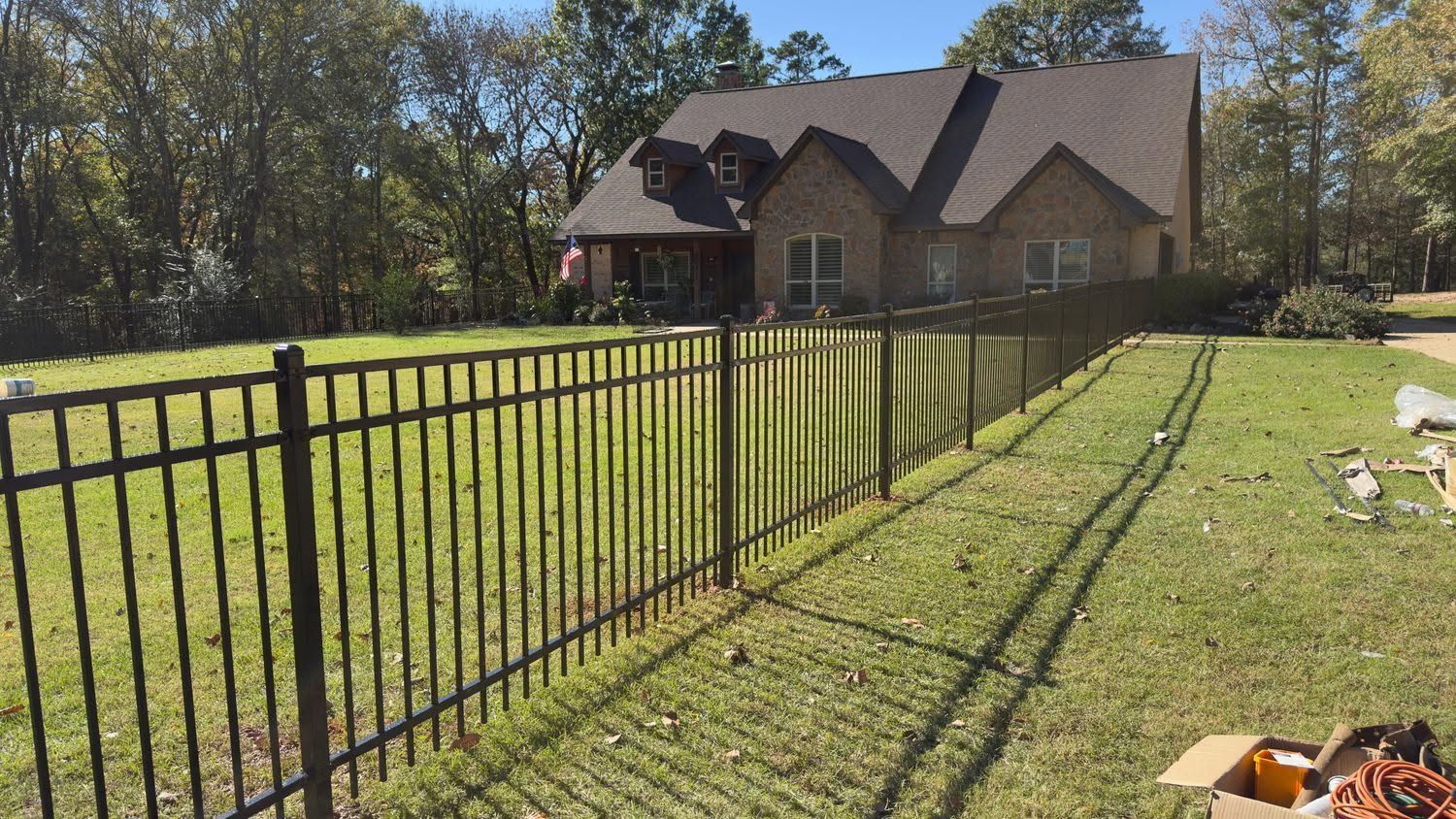 Black metal fence along a sunny grassy yard leading to a brick house with a covered porch.