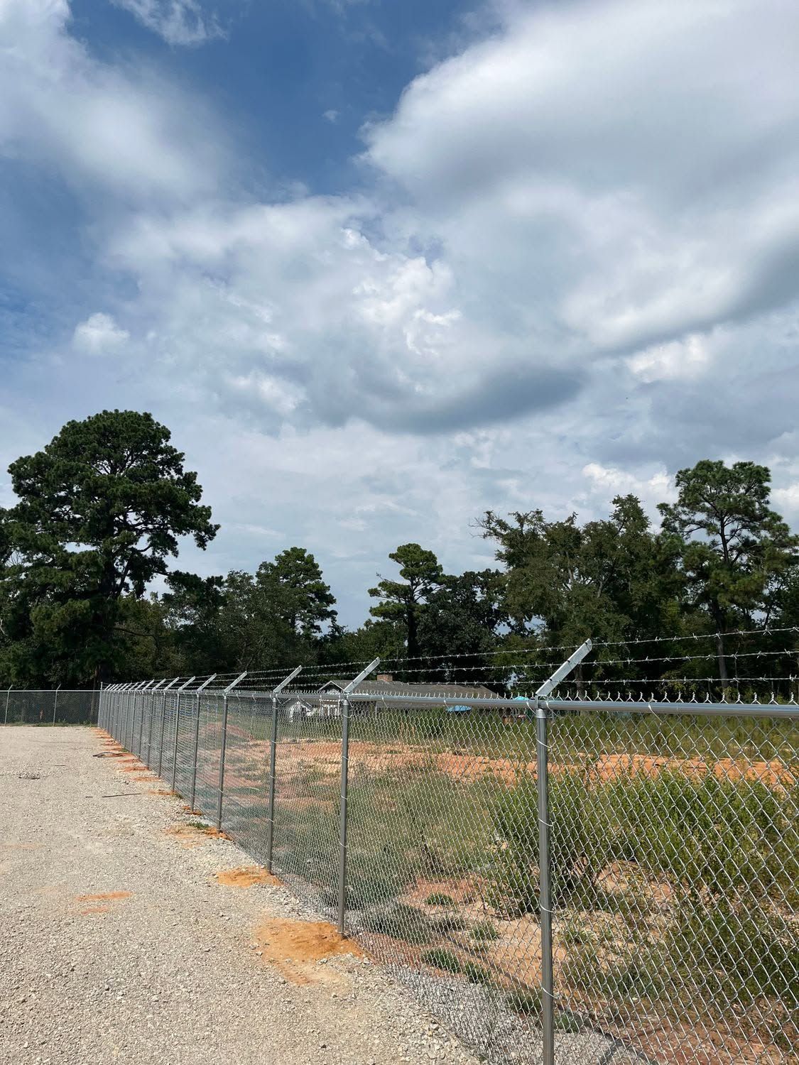 Chain-link fence bordering a gravel lot under a cloudy sky, with trees in the background.