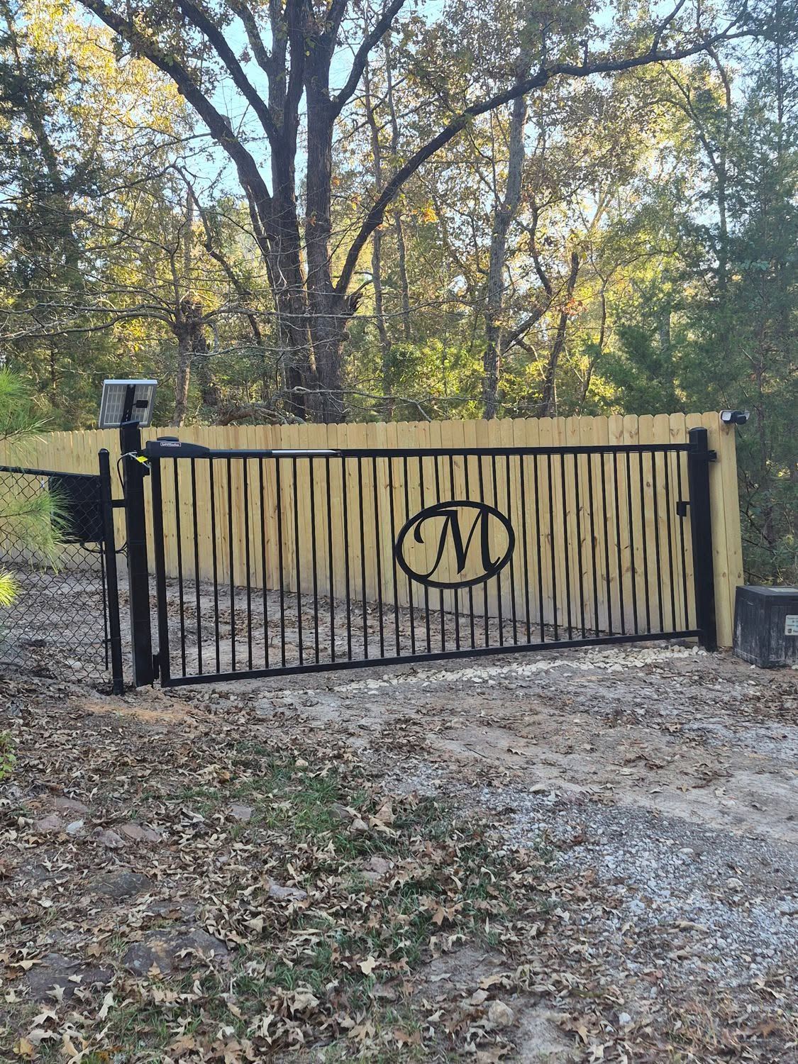 Black metal gate with circular monogram, set on a gravel driveway amid autumn trees.