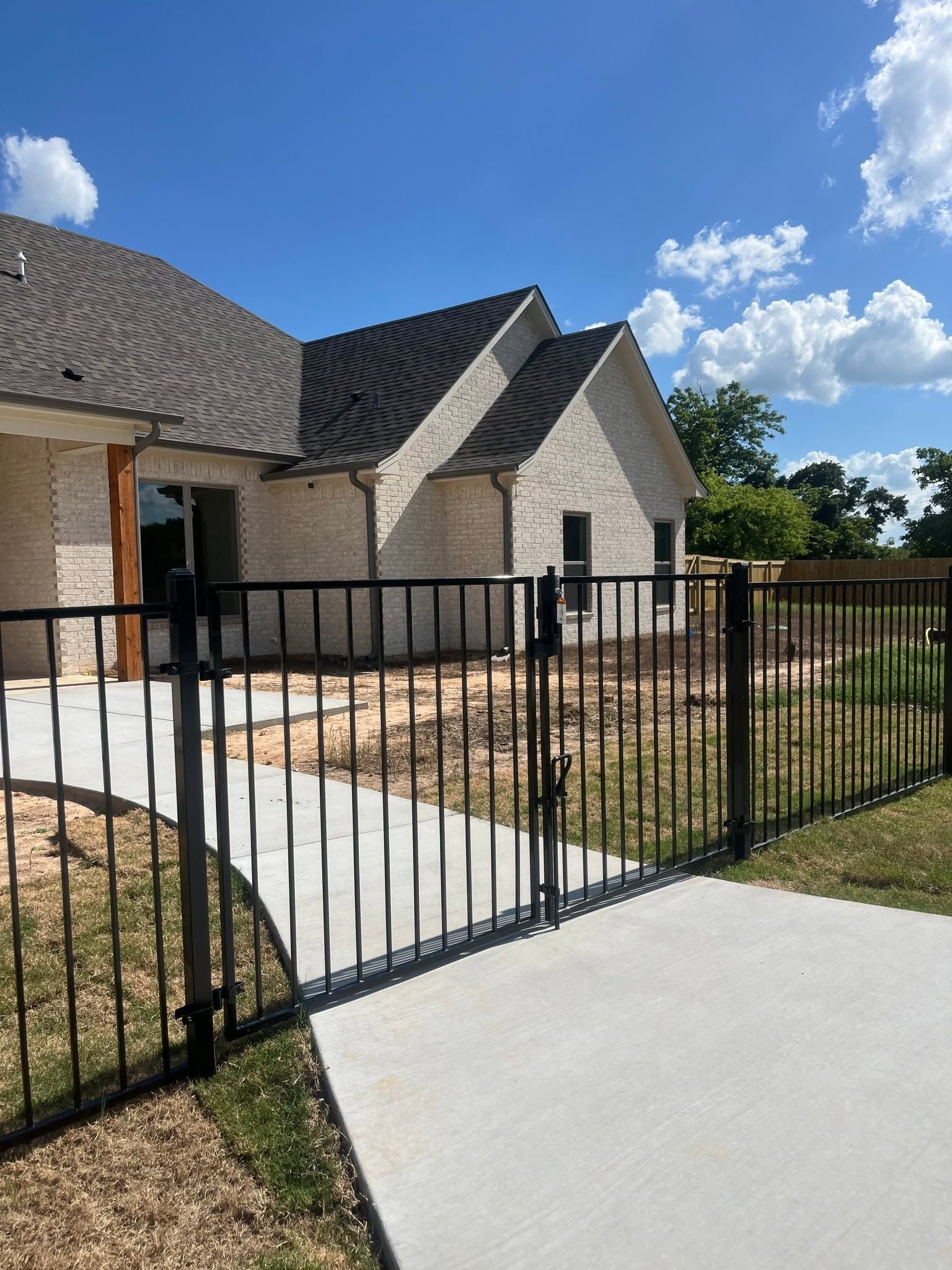 Black metal fence and concrete driveway in front of a brick house with a gray roof under a blue sky.