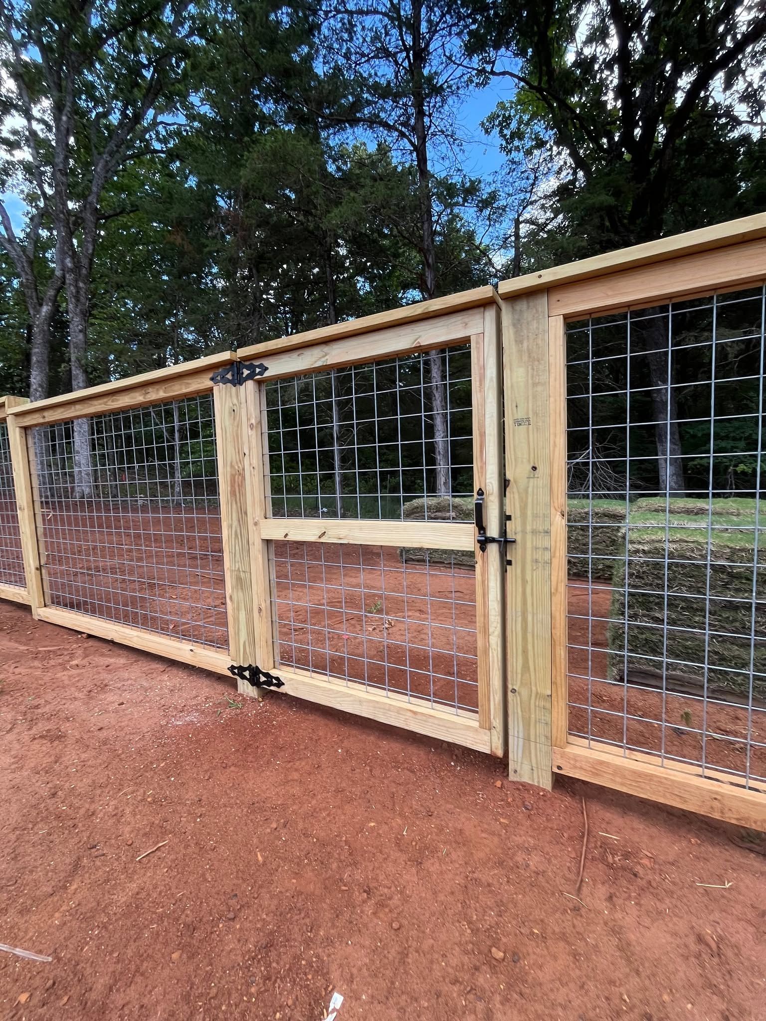 Wooden fence with wire panels and a gate along a red dirt path in a wooded area