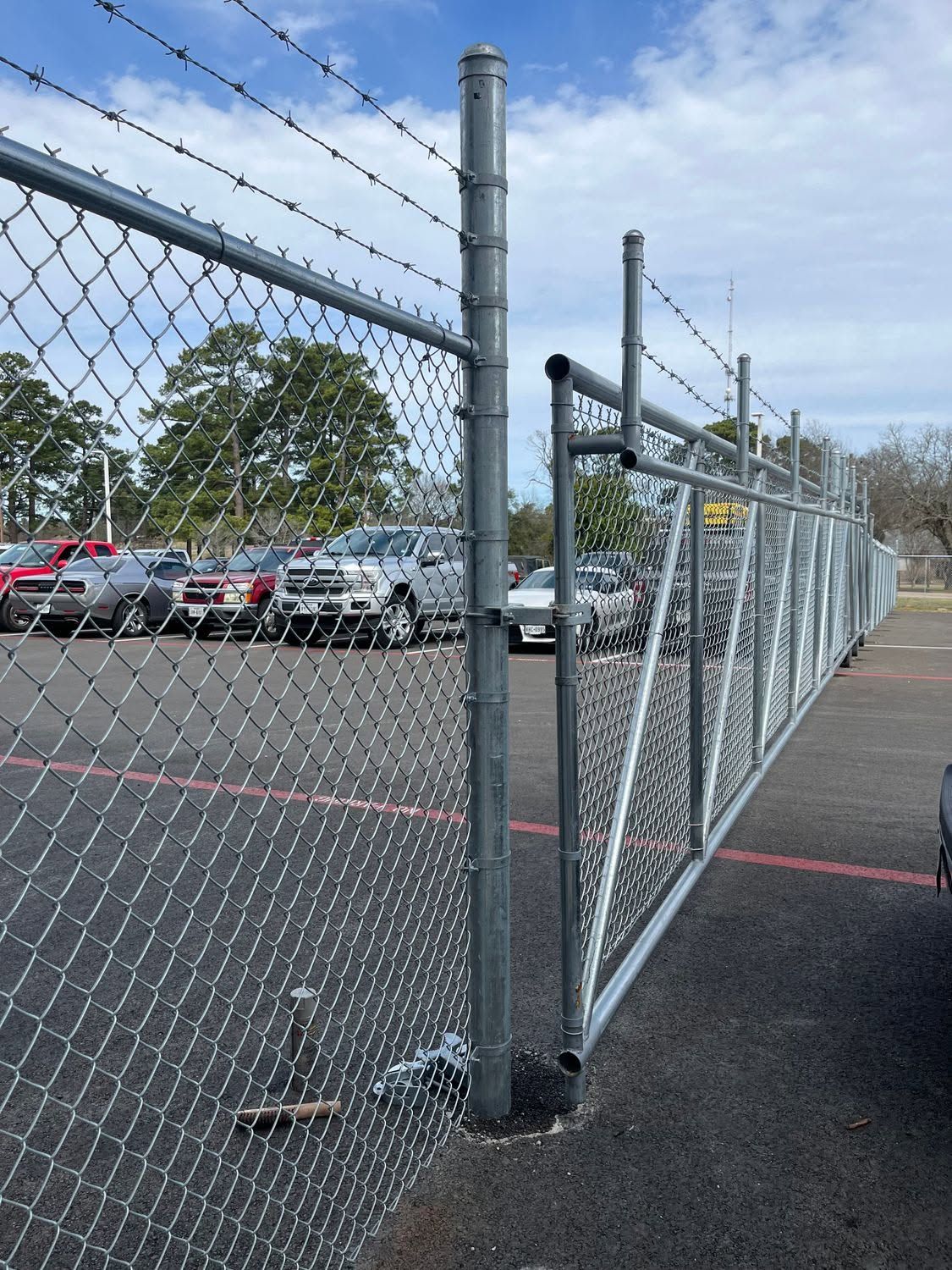 Chain-link security gate beside a parking lot, with barbed wire and red-marked pavement.