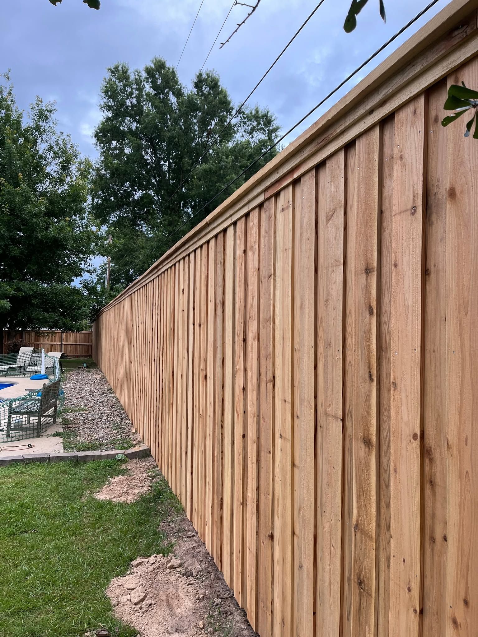 Tall wooden privacy fence along a backyard patio and grass, with trees and a pool in the background
