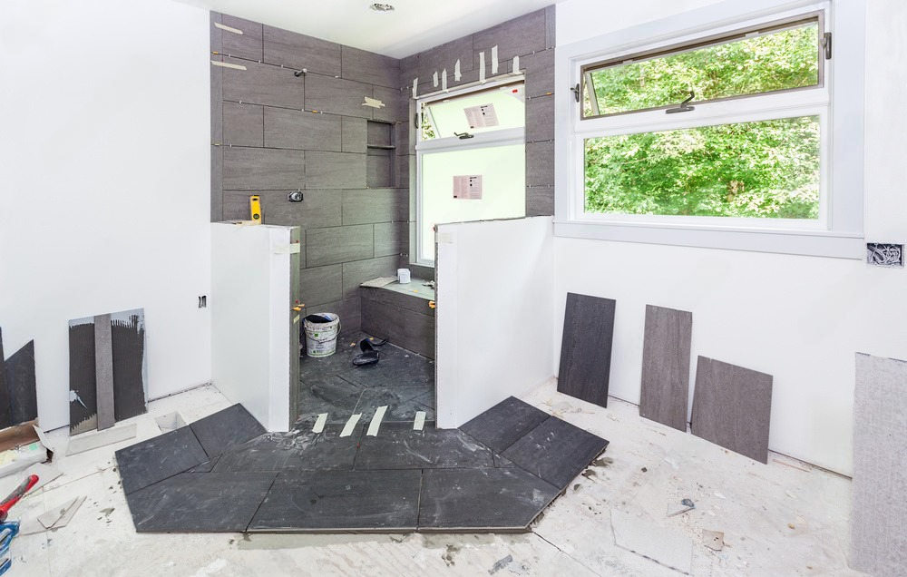 Bathroom under renovation with gray tiled walls and floor, white cabinets, and a window.