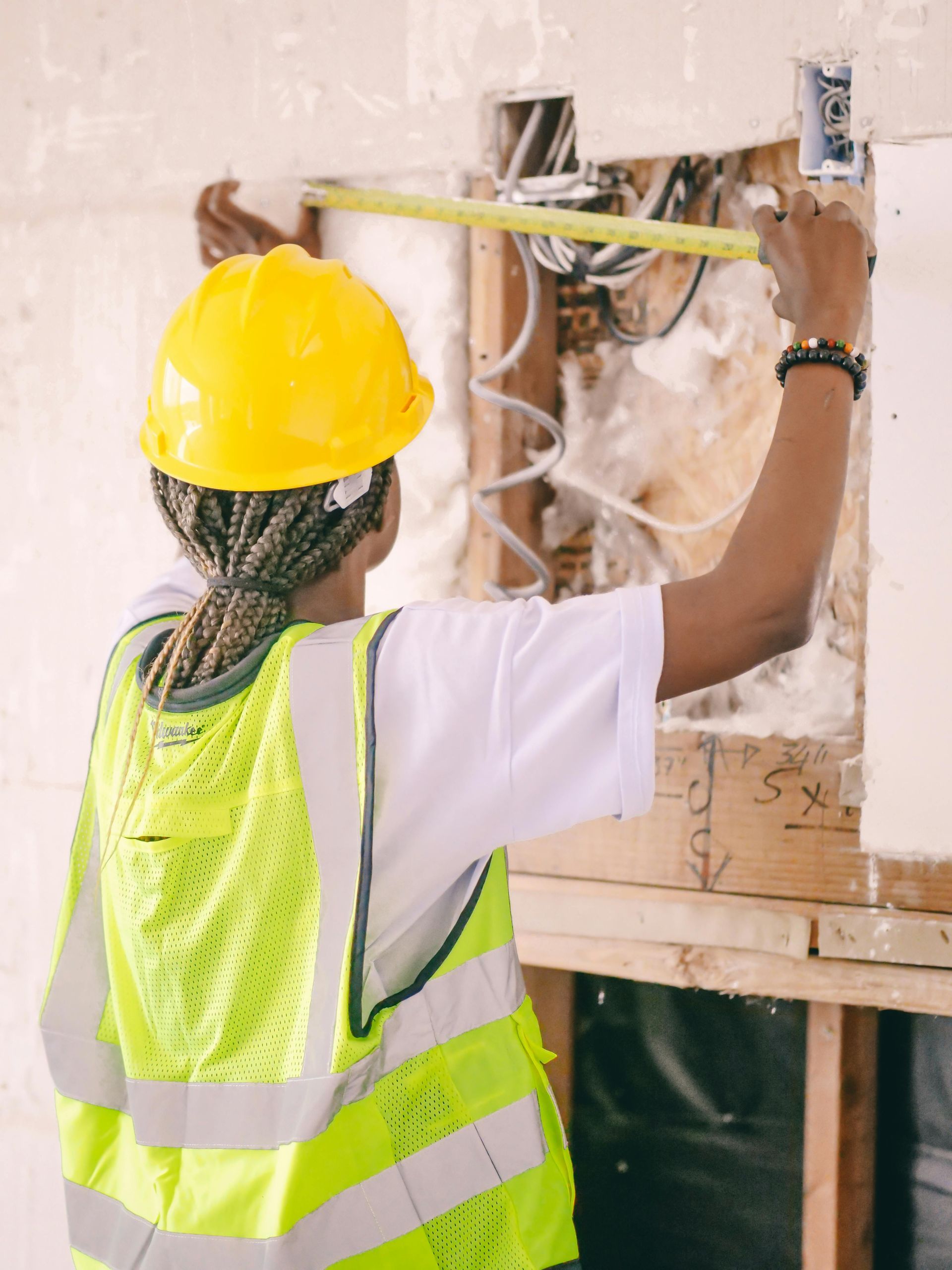 Person in hard hat and safety vest measuring an electrical box in a wall during construction.