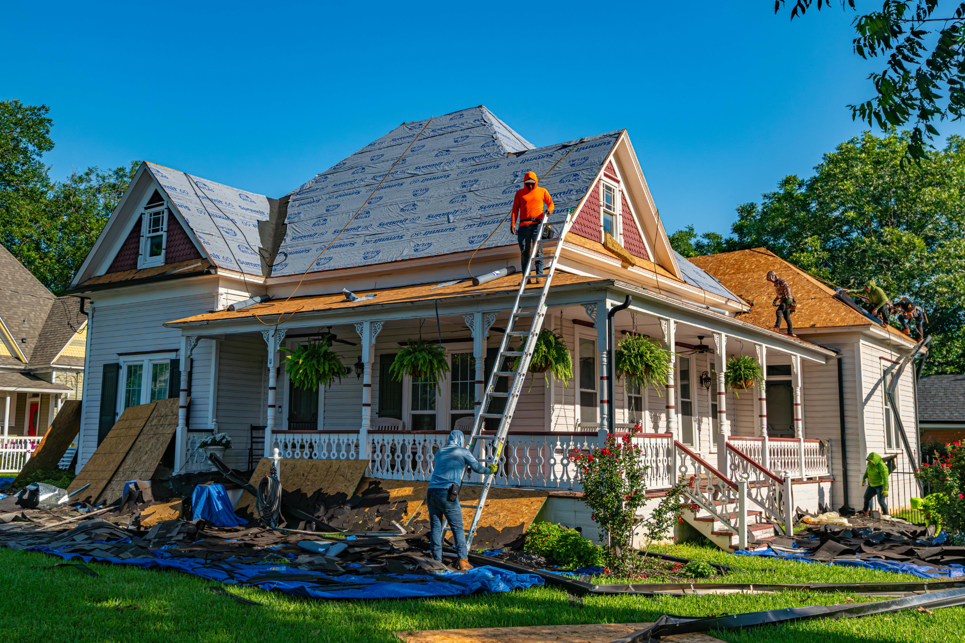 Workers repairing a house roof and porch under a clear blue sky, with ladders and construction materials nearby