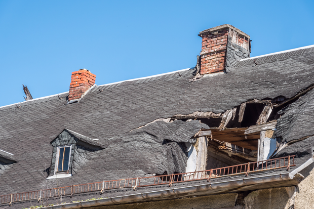 Damaged rooftop with a large collapsed section and exposed wooden beams under a blue sky