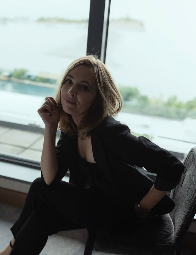 Woman in a dark outfit posing by a large window with a bright outdoor view.