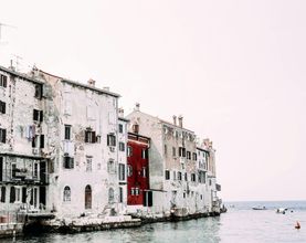 Weathered seaside buildings with a red facade beside calm water and small boats under a pale sky