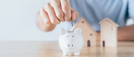 Hand placing a coin into a white piggy bank with small wooden houses in the background