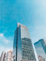 Tall modern glass skyscraper against a bright blue sky, with nearby buildings visible below.